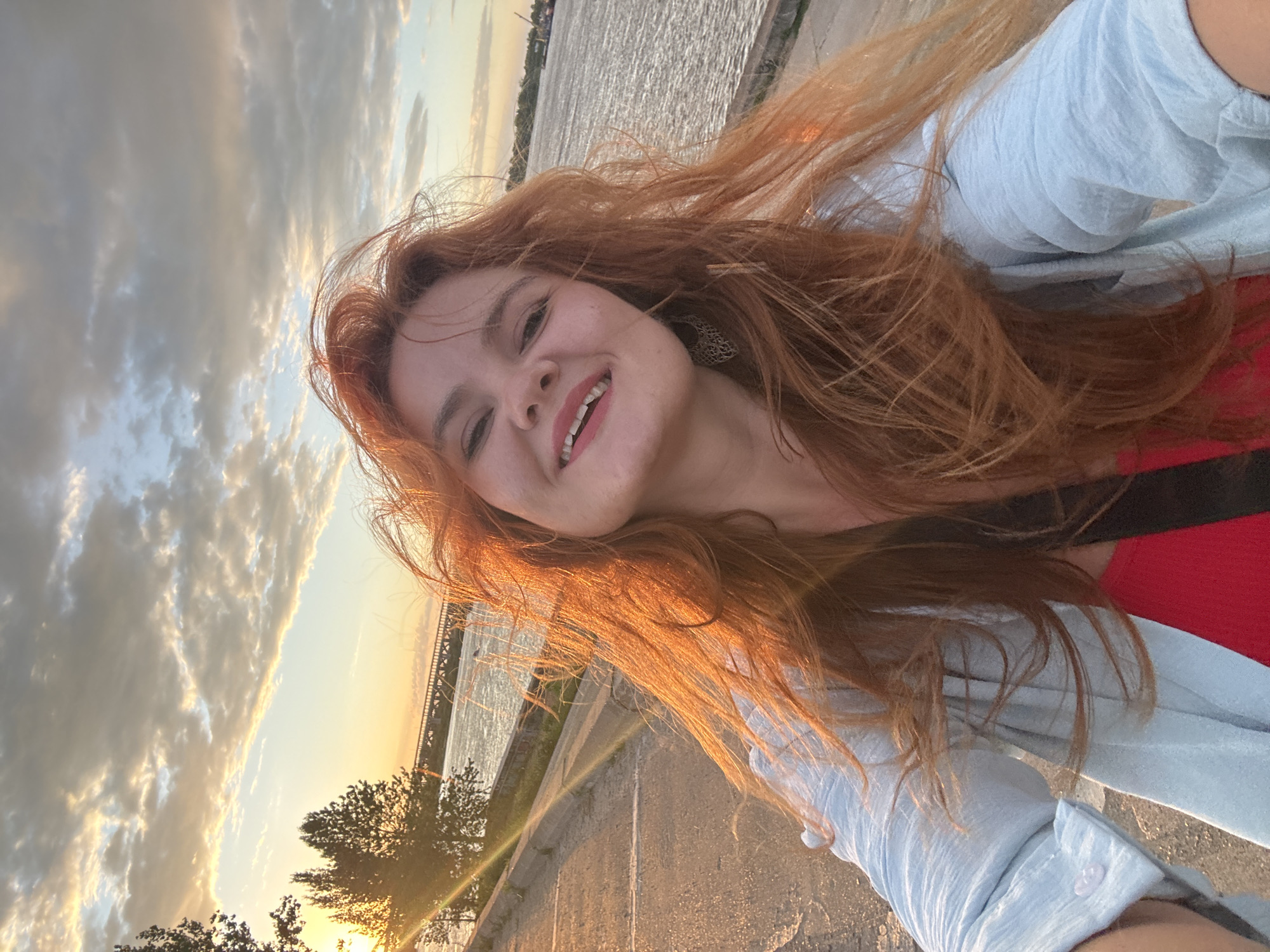 A woman with long, wavy red hair is taking a selfie outdoors. She is smiling and wearing a light blue shirt over a red top. She has a large, ornate earring in her left ear. The background shows a body of water, likely the Volga River, with a bridge in the distance. The sky is filled with dramatic clouds, illuminated by the setting sun, casting a warm glow on the scene. The location appears to be a waterfront promenade or embankment in Nizhny Novgorod, Russia, based on the presence of the Volga River and the characteristic architecture of the bridge. The lighting suggests it is late afternoon or early evening.