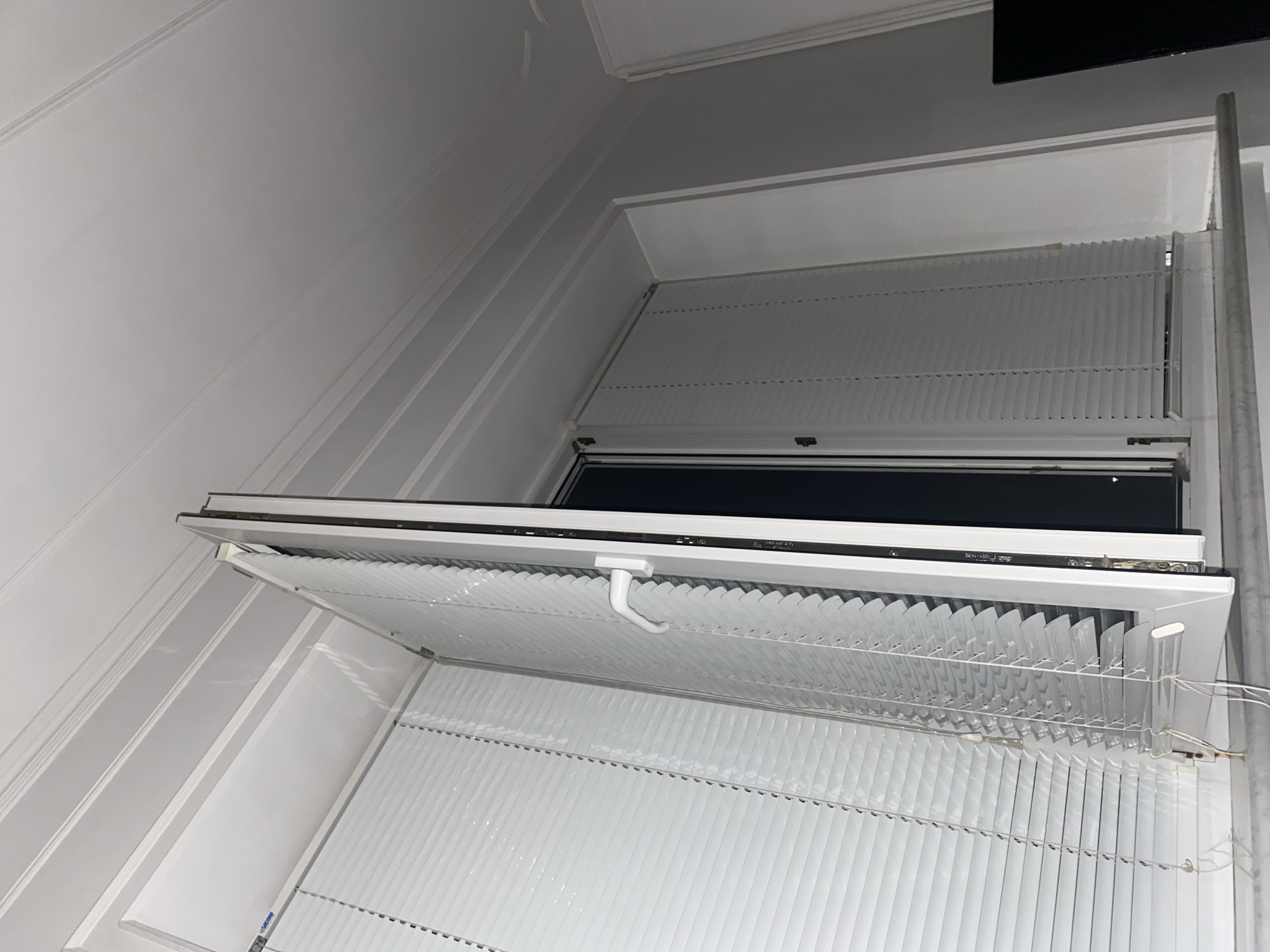 An interior shot shows a white-framed window with two sets of white horizontal blinds. The left window pane is closed, with its blinds fully down. The right window pane is open at an angle, revealing a dark interior. The blinds on the open pane are partially pulled up and angled. A white window handle is visible on the right pane. The scene appears to be a room with a white, paneled ceiling. There is no indication of the city of Suvorovskaya, Russia, or any specific environmental details that would suggest this location.