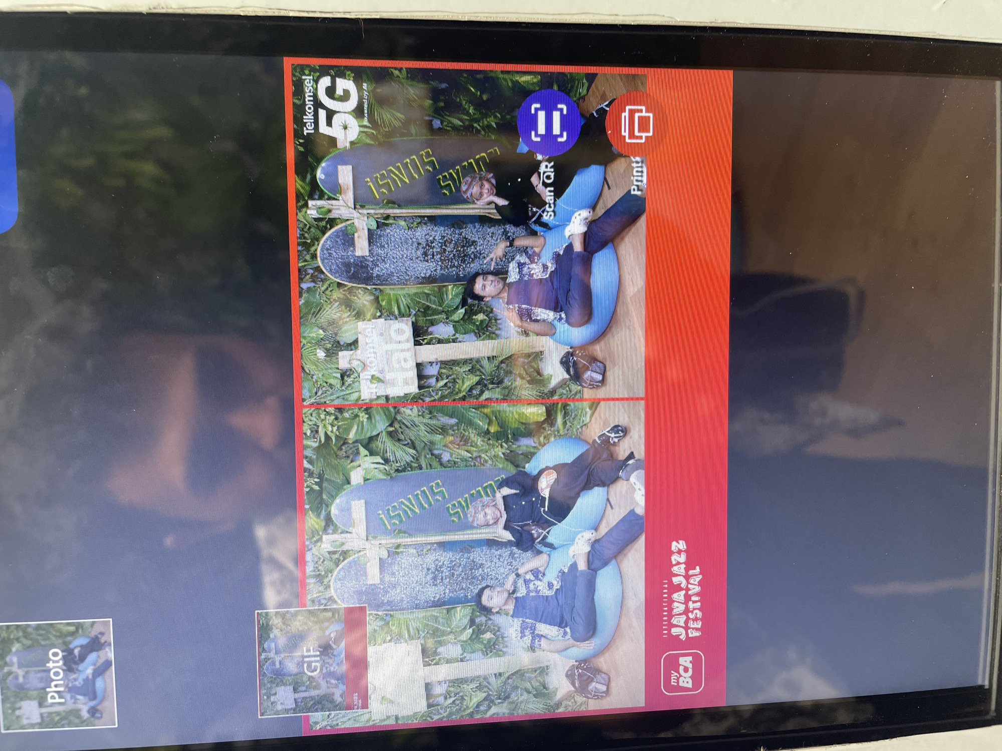 The image shows a digital display, likely a screen on a kiosk or similar device, displaying a photo collage. The upper right corner of the frame shows an advertisement for Telkomsel 5G. The main portion of the display contains what appears to be a photo print layout, featuring two identical images of two people (one man and one woman) posing in a decorated area. The location, as inferred from the text within the image, is Salembaran, Indonesia, as part of the Java Jazz Festival sponsored by BCA. The subjects are sitting on blue cushions with skateboards and foliage serving as a backdrop. There are options visible on the screen indicating features such as "Scan QR," "Print," and the option to create a GIF or a still "Photo." The image of the screen itself also includes a partial reflection of a face.