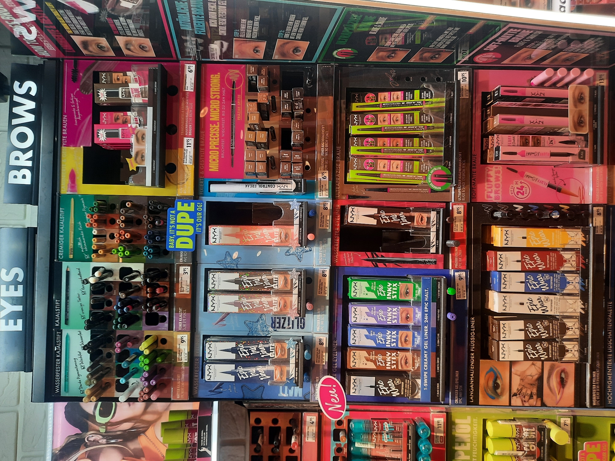 The image shows a retail display of NYX Professional Makeup products, specifically focusing on eye and brow makeup. The display is organized into sections labeled "EYES" and "BROWS". Various eyeliner pencils, liquid eyeliners, and brow pencils are showcased, with product names such as "Epic Ink liner," "Epic Wear," and "Inky Stix." The display features product packaging, testers, and descriptive text in German, indicating the location is a retail store. The products are arranged on shelves and in tiered displays. The lighting is bright, typical of a retail environment. The overall scene is a commercial product display. The store is likely located in Rosenheim, Germany, as indicated by the German text on the product packaging and signage.
