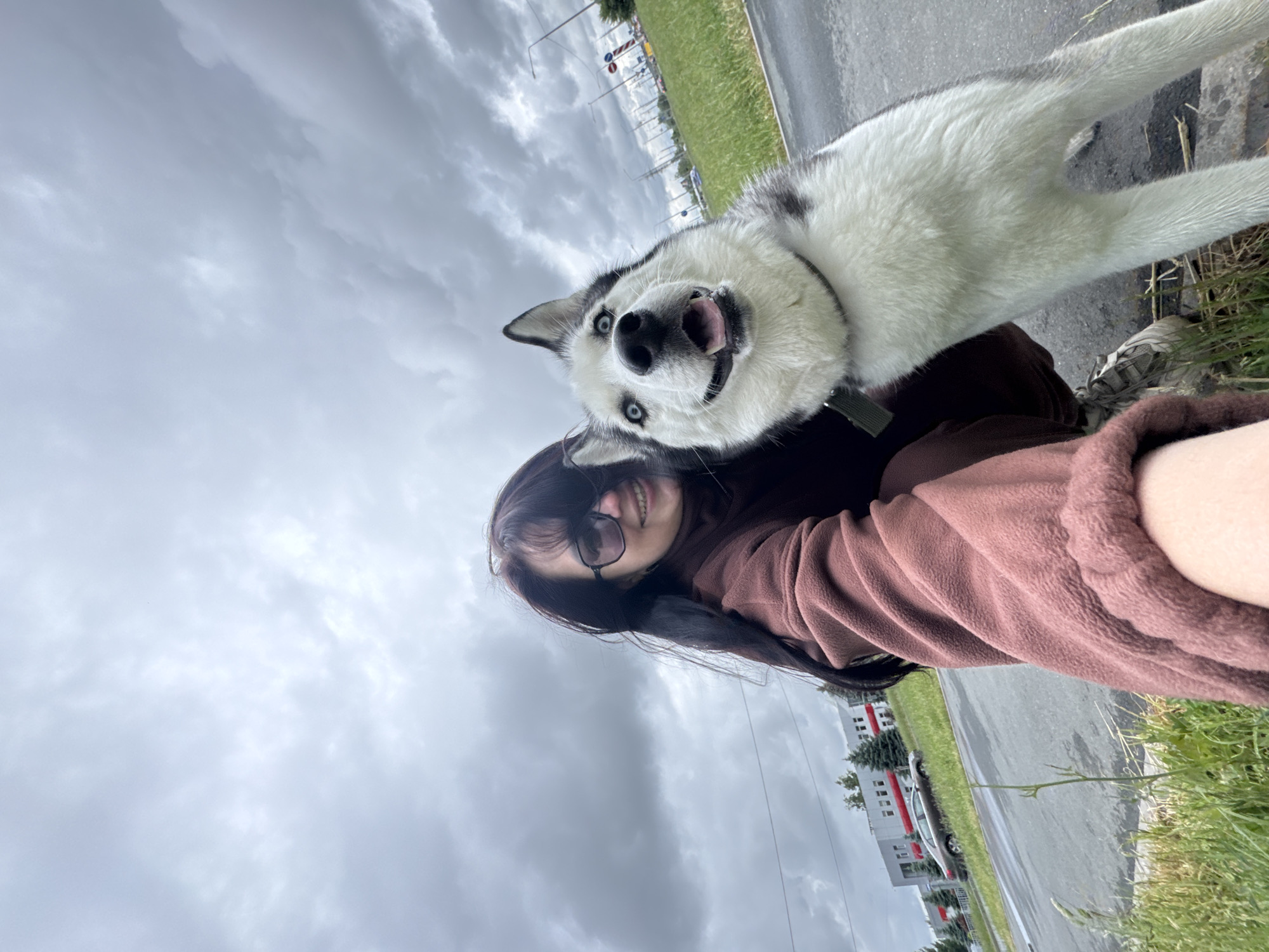 A woman with long, dark hair and sunglasses is shown from the chest up, holding a white and black husky dog with blue eyes. Both subjects face the camera with a slightly tilted angle. The dog's mouth is open, appearing playful. The woman wears a maroon fleece jacket. They are standing next to a sidewalk and street, with overcast skies above. Behind them are buildings and greenery, suggesting a residential or commercial area. The location is in the city of Unknown, Unknown.
