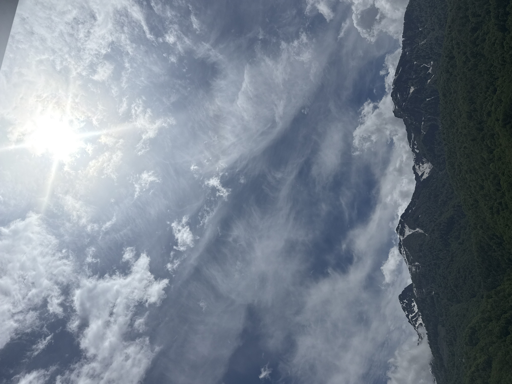 The image depicts a mountain landscape in Krasnaya Polyana, Russia. The sky is predominantly visible, featuring a mix of white cumulus clouds and thin cirrus clouds. The sun shines brightly through a break in the clouds, creating a lens flare effect. A mountain range covered in dense green forest forms the right edge of the frame. The mountain face exhibits patches of snow and bare rock.
