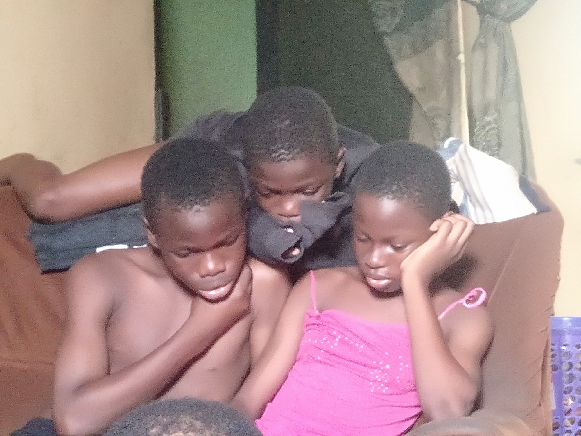 The photograph depicts three young children, two boys and one girl, seated closely together on a brown upholstered sofa, likely within a home in Shangisha, Nigeria. All three children have dark skin and short, dark hair. The boy on the left is bare-chested, his right hand resting near his chin, as he looks intently downwards. The central figure, another boy, is wearing a dark top and appears to be leaning forward, also focused downwards. On the right, a girl in a vibrant pink, sequined strappy top supports her head with her left hand, her gaze also directed downwards. Their unified focus suggests they are engrossed in a shared activity, possibly watching a screen or reading something together, though the object of their attention is not visible.
The background shows interior walls, with a light beige or cream color on the left and a darker green hue on the right. A window covered by light-colored curtains is discernible in the upper right quadrant. A striped fabric, possibly a pillow or blanket, is draped behind the children. On the far right, a portion of a purple plastic basket or container is visible. The lighting in the scene is somewhat subdued, contributing to a calm atmosphere, and the image itself exhibits a slightly grainy texture. The children appear calm and concentrated during what seems to be a moment of leisure. No text is visible in the image.
