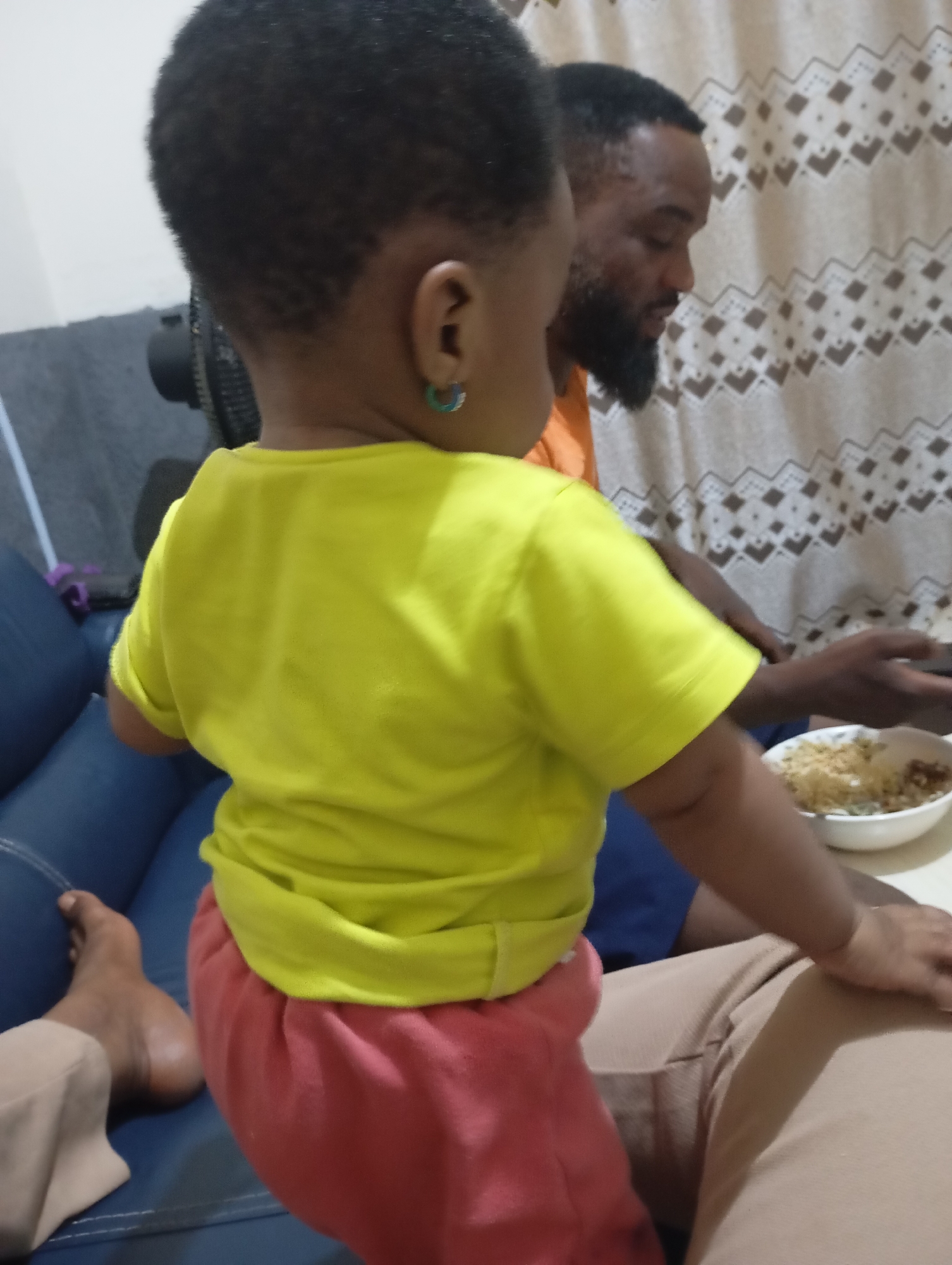 In a dimly lit indoor setting, possibly a living room in New Stadium, Nigeria, a young child with dark, short hair and wearing a bright yellow t-shirt and pink pants is positioned in the foreground. The child has a green earring in their left ear. Behind the child, an adult male with dark hair and a beard, wearing an orange shirt, is visible. The man appears to be looking down at a bowl of food. The background features a patterned curtain and furniture, including a blue sofa. A fan is partially visible behind the child. The overall atmosphere suggests a casual family gathering.