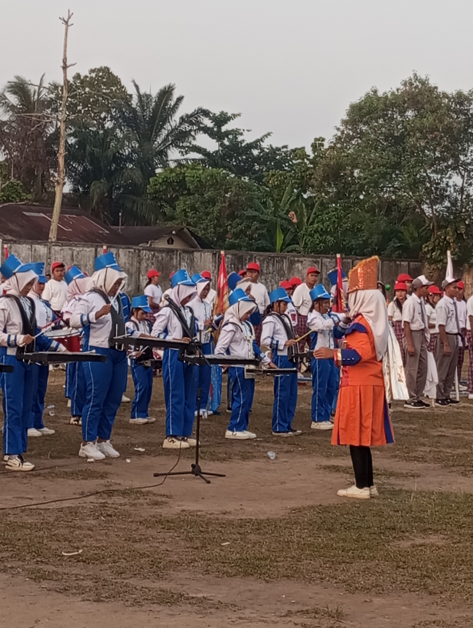A marching band, composed of young individuals in blue and white uniforms with blue hats and headscarves, stands in formation on a dirt and grass field. The band members are holding musical instruments, some appearing to be drums and xylophones. A person in a prominent orange and blue uniform, possibly a conductor, faces the band. In the background, a concrete wall, trees, and buildings are visible under an overcast sky. The scene is likely taking place in Tanjung Gading, Indonesia, given the context provided.