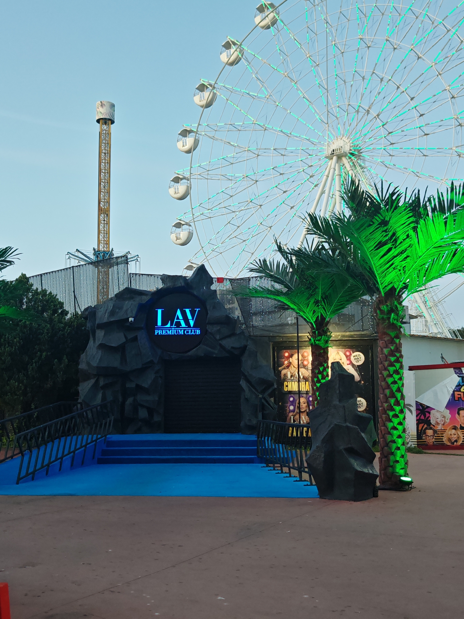 The image depicts the entrance to LAV Premium Club in Nesebar, Bulgaria, situated within a park setting with amusement park rides in the background. The entrance is designed to resemble a dark, artificial cave, with a blue illuminated sign displaying "LAV PREMIUM CLUB." Blue carpeting extends up the stairs leading into the club's closed entrance. Adjacent to the entrance are artificial palm trees lit with green lights. Posters advertising the club's events are visible on the walls flanking the entrance. In the background, a large Ferris wheel with green lights is partially visible, along with a tall amusement park drop tower. A black metal railing lines the side of the entrance stairs. The ground in front of the entrance is paved and appears to be damp. The sky is pale blue.