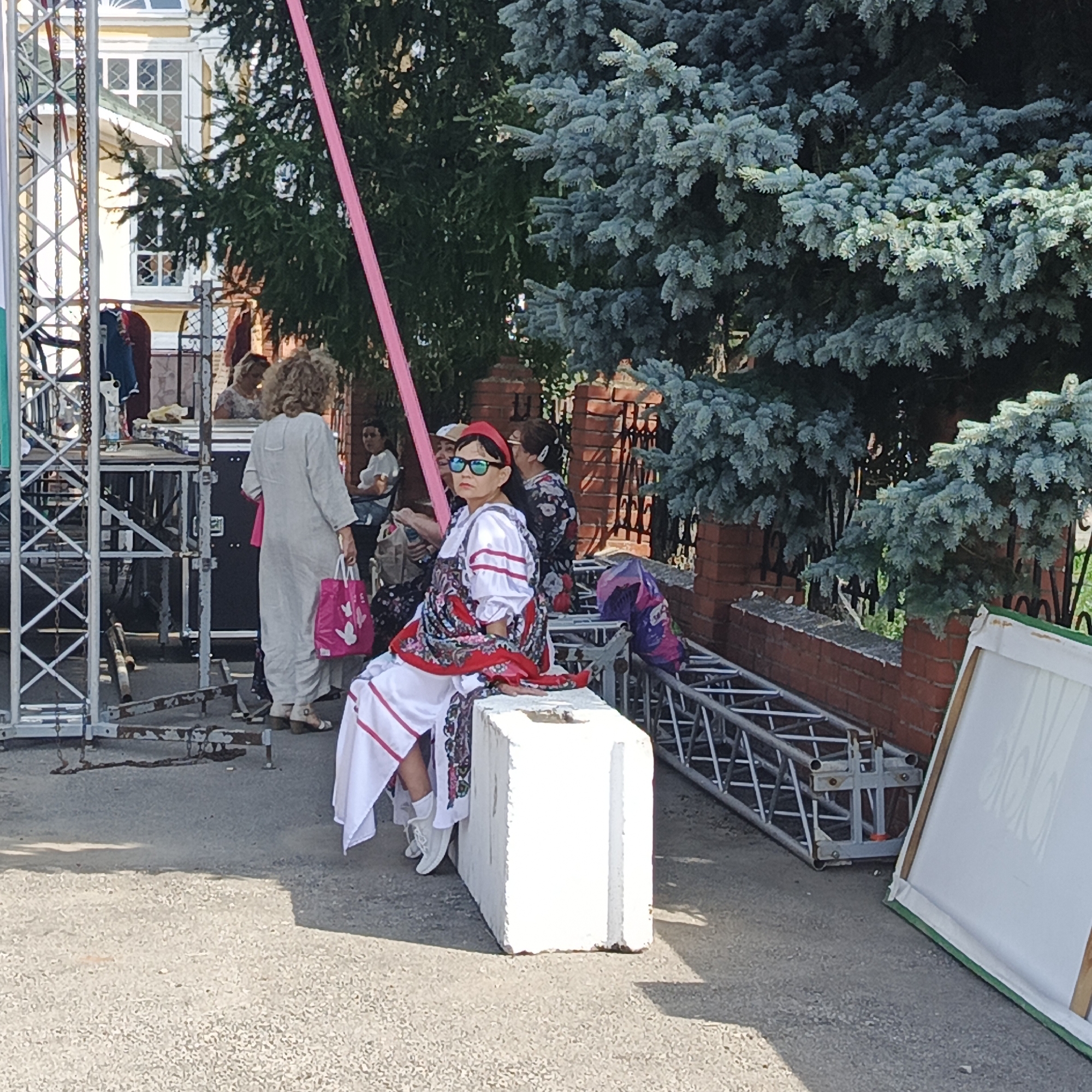 In Nikolo-Beryozovka, Russia, a young woman in traditional attire sits on a concrete block, facing right. She wears a red headband, sunglasses, a white dress with red and black embroidery, and white shoes. Her hands rest on her lap, and she is looking slightly down. Behind her, other people are visible in the background, along with scaffolding, trees, and a brick wall. The scene appears to be outdoors, possibly at a festival or event.