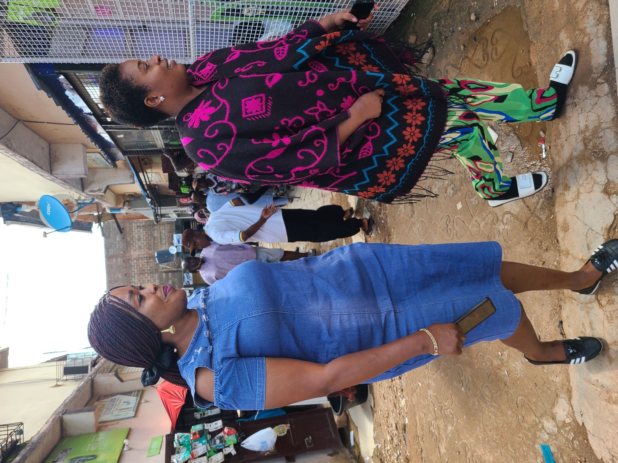The image shows two women standing on a street in Jos, Nigeria. The woman in the foreground is wearing a blue denim dress and black Adidas slides. She has dark braided hair and is wearing gold earrings and a bracelet. She is holding a black smartphone. The woman in the background is wearing a colorful patterned outfit and a black and pink patterned shawl. She is holding a black smartphone. Several other people are visible in the background, as well as buildings and market stalls. The ground is paved with concrete and dirt, and there are puddles of water, suggesting recent rain. The scene appears to be a busy street market. The time of day is difficult to determine, but the lighting suggests it is daytime. There is no visible text in the image.