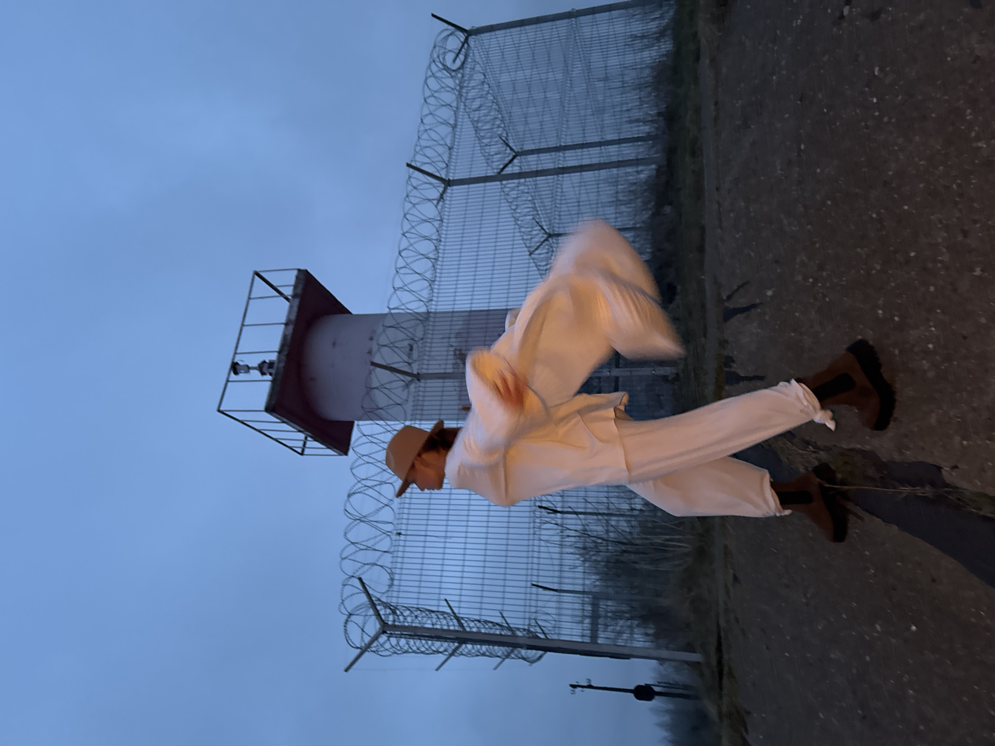 An outdoor, vertically oriented photograph captures a single individual in a dynamic pose against a muted, overcast sky. The person, positioned in the middle ground, wears a light brown fedora, a white long-sleeved top or jacket, white trousers, and dark brown boots. Their body is in motion, with one leg raised and arms extended, creating a blur of white fabric or an object near their right arm, suggesting an active movement like dancing, leaping, or performing.
In the background, a tall, slender structure resembling a lighthouse or a communication tower, with a red top and a light-colored body, rises towards the sky. A prominent chain-link fence, topped with multiple strands of barbed or razor wire, runs horizontally behind the person, spanning across the mid-section of the image. Beyond the fence, the silhouettes of bare trees or sparse vegetation are faintly visible against the dark, featureless sky, indicating a natural or rural setting in Chaikovsky, Russia. The foreground consists of a dark, paved or asphalt surface.
The overall lighting suggests either dusk or dawn due to the dim, cool blue and gray tones of the sky. The combination of the fence with razor wire and the tower implies a possibly restricted or industrial area. No text is visible in the image. The scene conveys a stark and somewhat dramatic atmosphere.