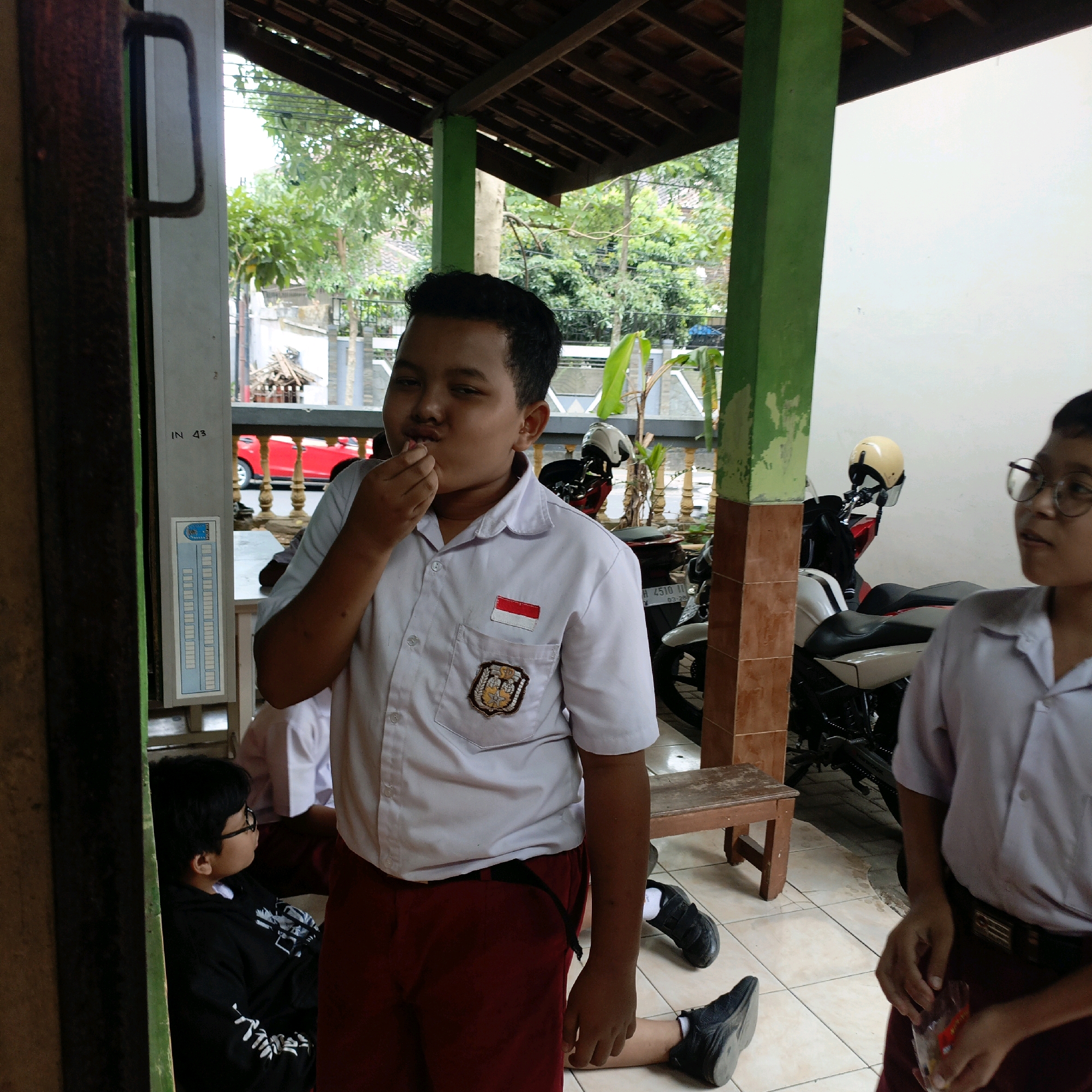 The image captures a candid moment at what appears to be an outdoor educational facility or gathering place, likely within the city of Salatiga, Indonesia. In the foreground, a young boy wearing a white school uniform with red shorts is in the center, bringing something to his mouth with his right hand. His uniform features an Indonesian flag patch on the left chest and an emblem on the right breast pocket. To his left, another young boy in a black hoodie with white designs and glasses sits on the ground, facing away from the camera. Partially visible in the background are two motorcycles parked near a white wall. A wooden bench is positioned between the boys and the motorcycles. The environment is partially covered by a wooden-slatted roof supported by green and brown posts. Trees and other buildings are visible in the distance, suggesting a semi-urban or suburban setting. The overall impression is one of a relaxed, possibly school-related, outdoor break.