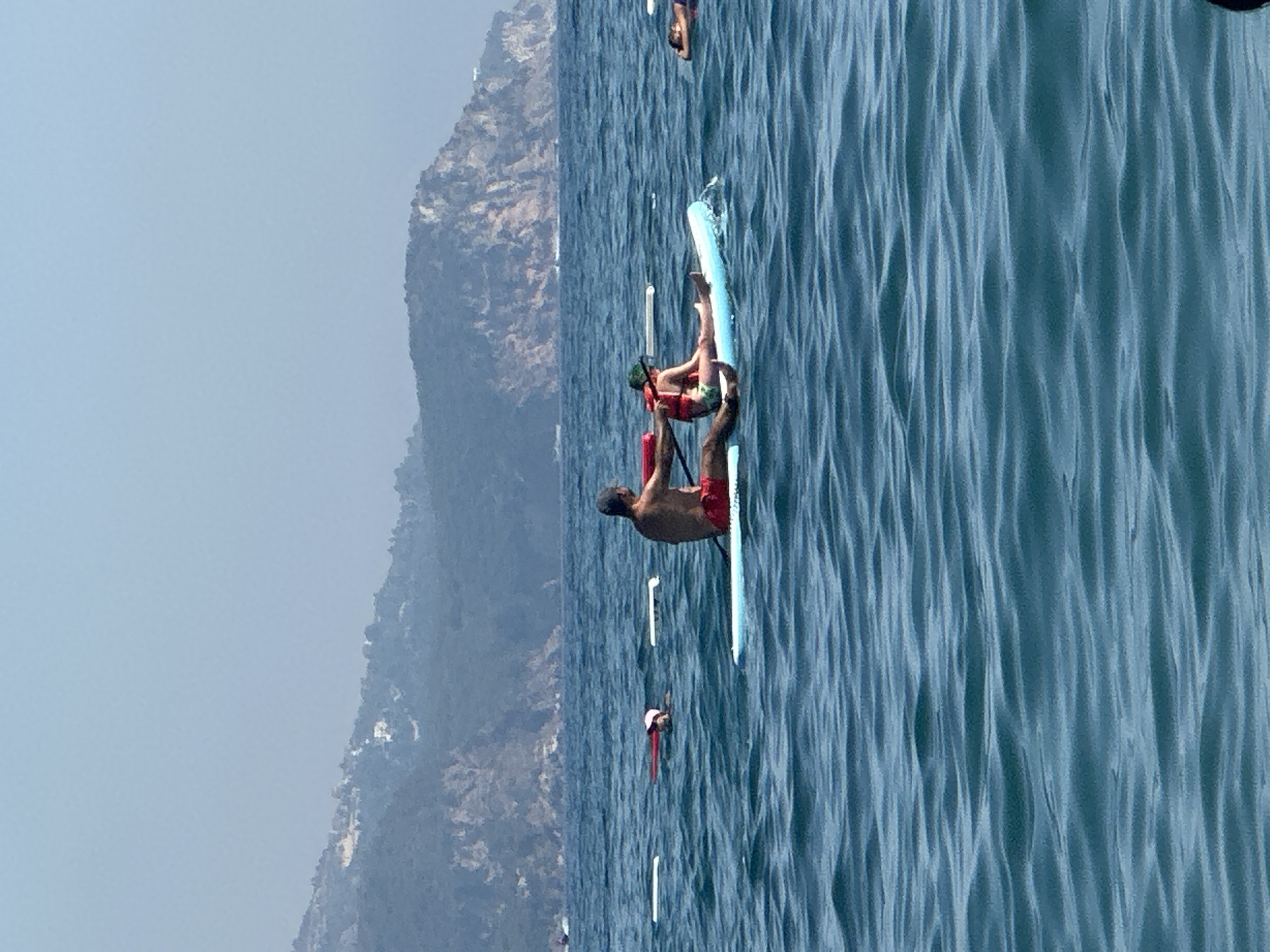 The image shows a group of people on paddleboards in the sea. In the foreground, a man and a child are on a paddleboard. The man is paddling and the child is sitting. Both are wearing life vests. The child is wearing a green hat. In the background, there are mountains and a hazy sky. There are also other people on paddleboards in the distance. The water is blue and there are waves. The scene appears to be in Boreti, Montenegro, a coastal town known for its beaches and clear waters.