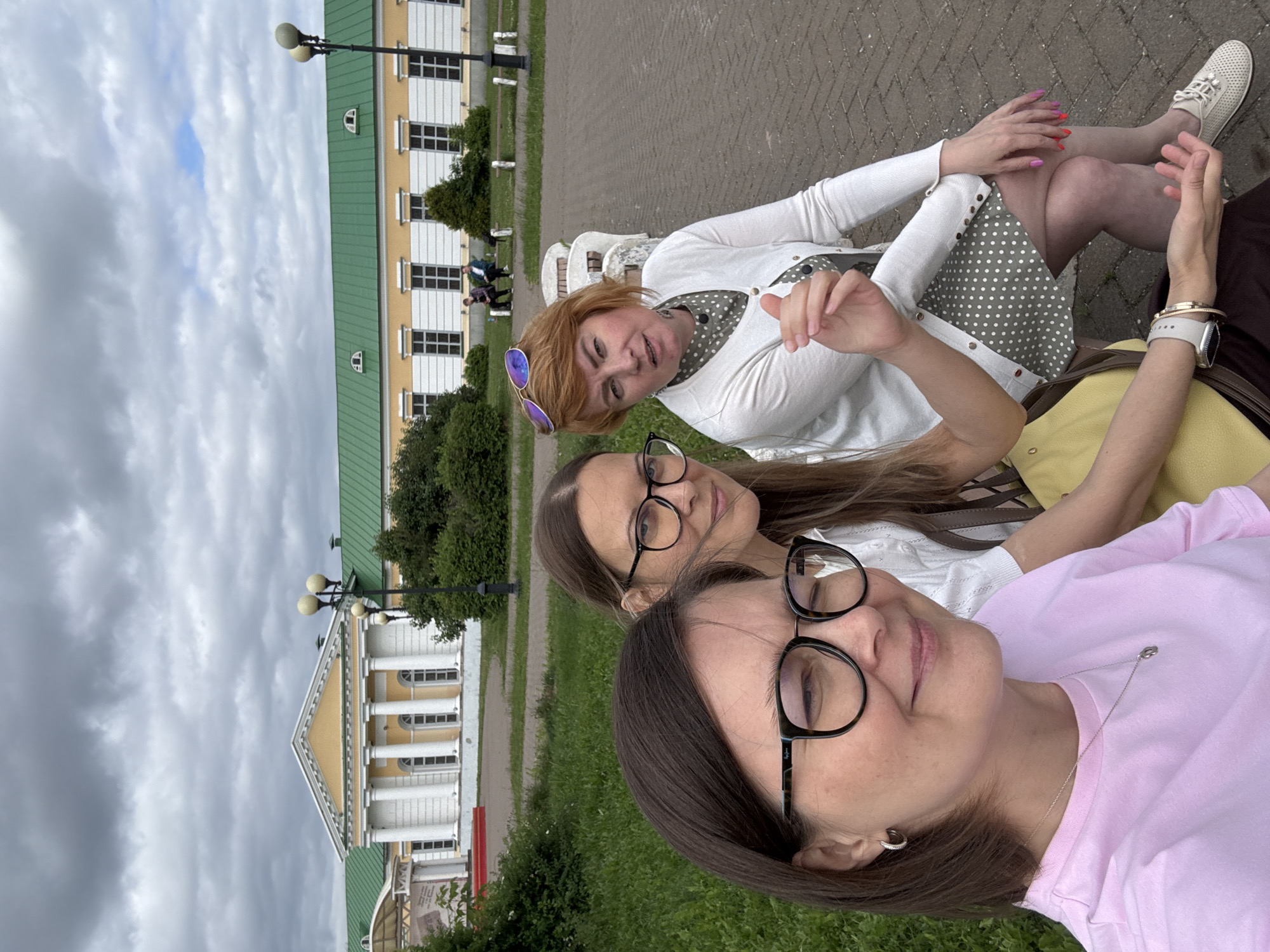 Three women are posing for a selfie on a park bench in front of a historical building. The building, with a yellow facade and a green roof, has a classical architectural style with columns and arched windows. The women are dressed casually, with one wearing a pink t-shirt, another a white blouse with a polka-dot dress and cardigan, and the third a white blouse. They are smiling and appear to be enjoying their outing. The background shows a well-maintained park with green grass, trees, and paved walkways. The sky is cloudy. The building in the background is likely part of the National Museum of the Udmurt Republic, formerly known as the residence of the P. I. Chaikovsky.
