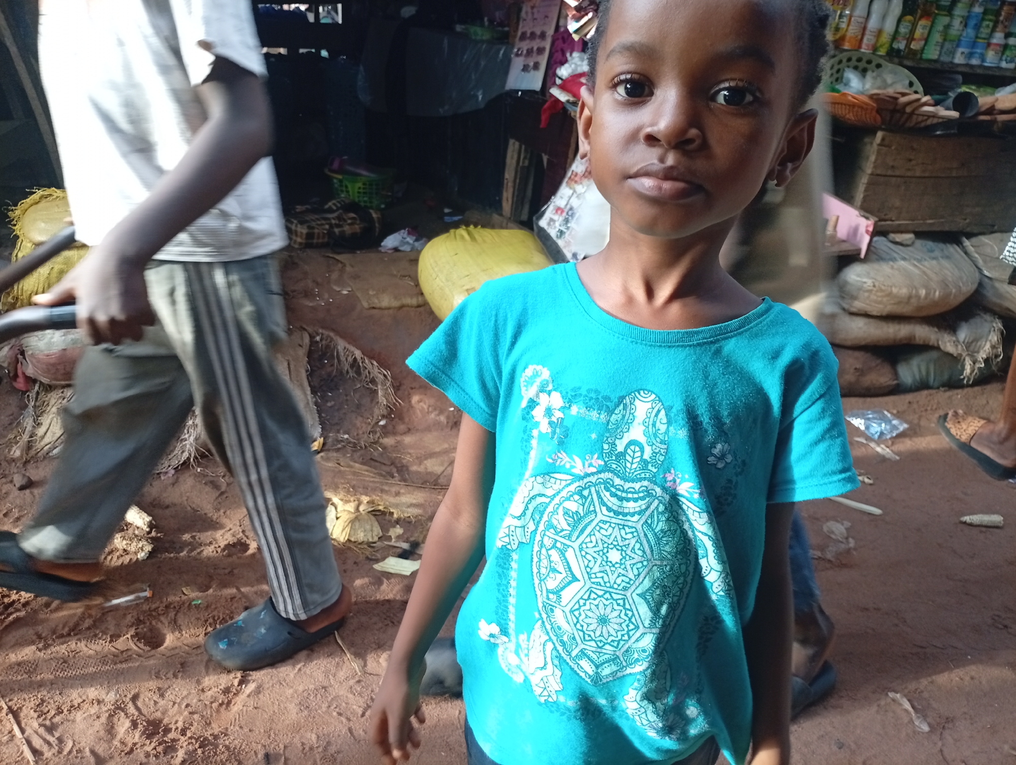 In Nkpor, Nigeria, a young Black girl with dark skin and large eyes stands front and center, looking directly at the camera. She wears a bright teal t-shirt adorned with a white turtle graphic. To her left, a person in a white t-shirt and grey athletic pants with white stripes walks away, their dark clogs visible on the sandy ground. Behind the girl, the scene appears to be a market or cluttered outdoor space with wooden structures, shelves stocked with items, and various goods piled on the ground, including large yellow and brown sacks, and a green basket. The ground is a mixture of sand and dirt, with scattered debris.