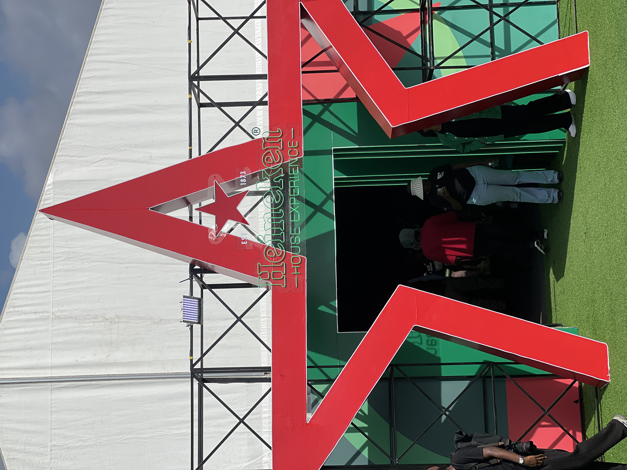 The image shows the Heineken House Experience in Lagos, Nigeria. A large, red Heineken star logo is prominently displayed against a green and white backdrop, with people entering the building. The environment appears to be an outdoor event space. A security guard is lying on the ground in the lower right corner.