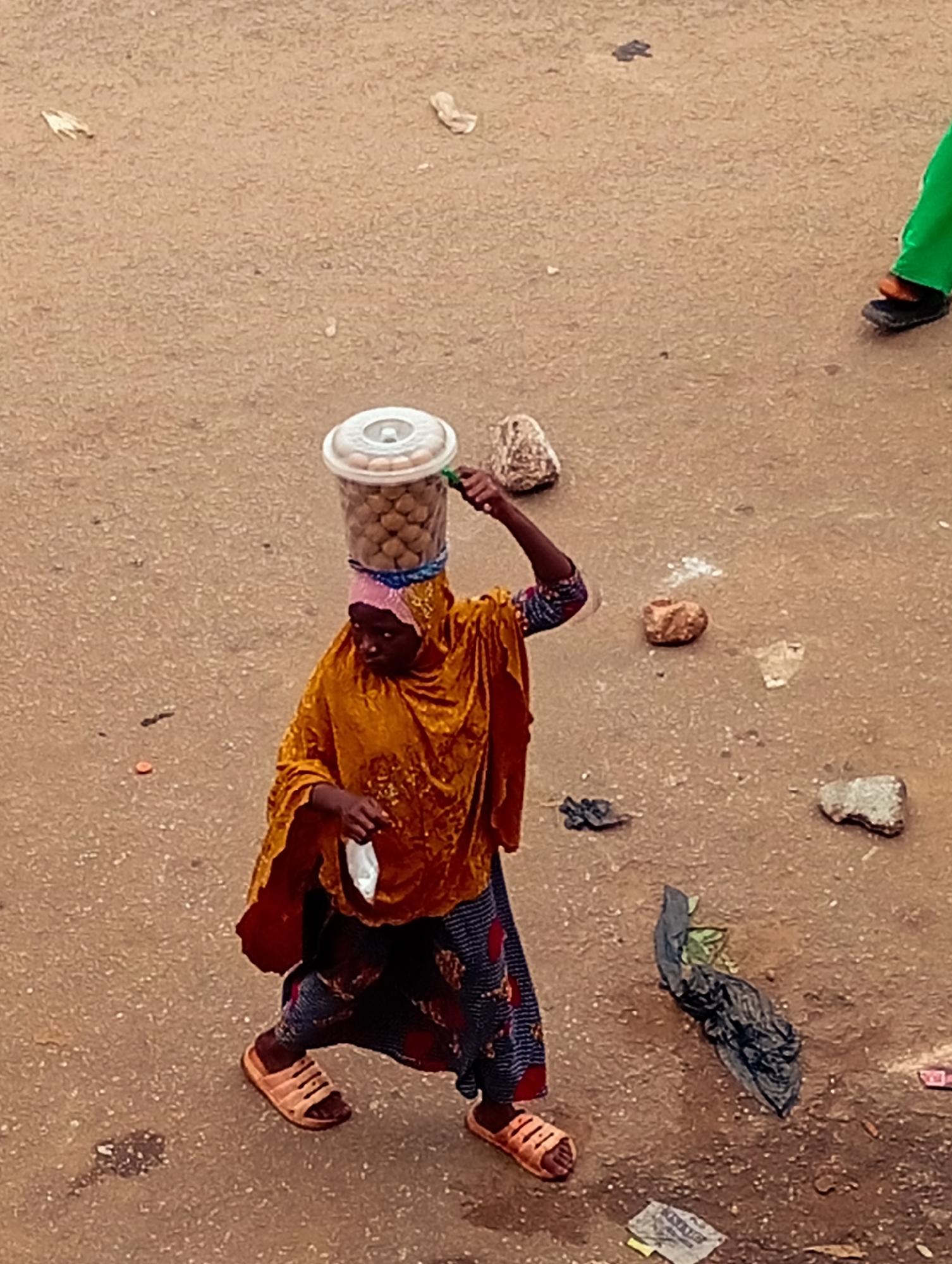 In Bukuru, Nigeria, a woman walks on a dirt road, carrying a clear plastic container filled with small, round, brown items on her head. She is wearing a vibrant orange, shimmering headscarf and a matching long-sleeved garment draped over her shoulders. Her lower body is clad in a patterned blue and red skirt, and she wears open-toed tan sandals. In her right hand, she holds a white object, possibly a cloth or bag. The ground around her is predominantly a reddish-brown dirt, scattered with small rocks, debris, and a crumpled black plastic bag. To the right, another person's legs are visible, wearing green pants and dark sandals.