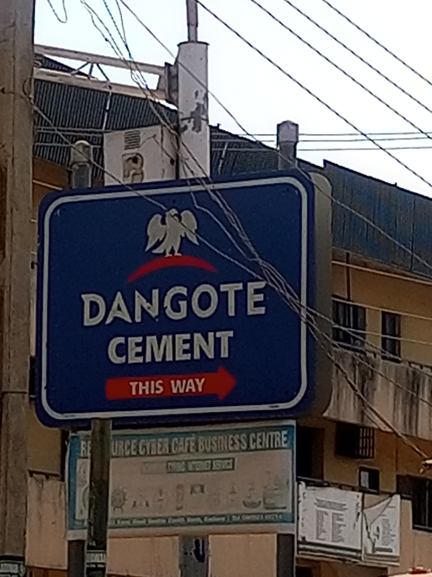 The image shows a street in Maiburiji, Nigeria. A blue and white rectangular sign for Dangote Cement points to the right. Below it is another sign advertising the "URCE Cyber Cafe Business Centre." The signs are mounted on a pole with electrical wires obscuring some of the view. A building with barred windows is visible in the background.