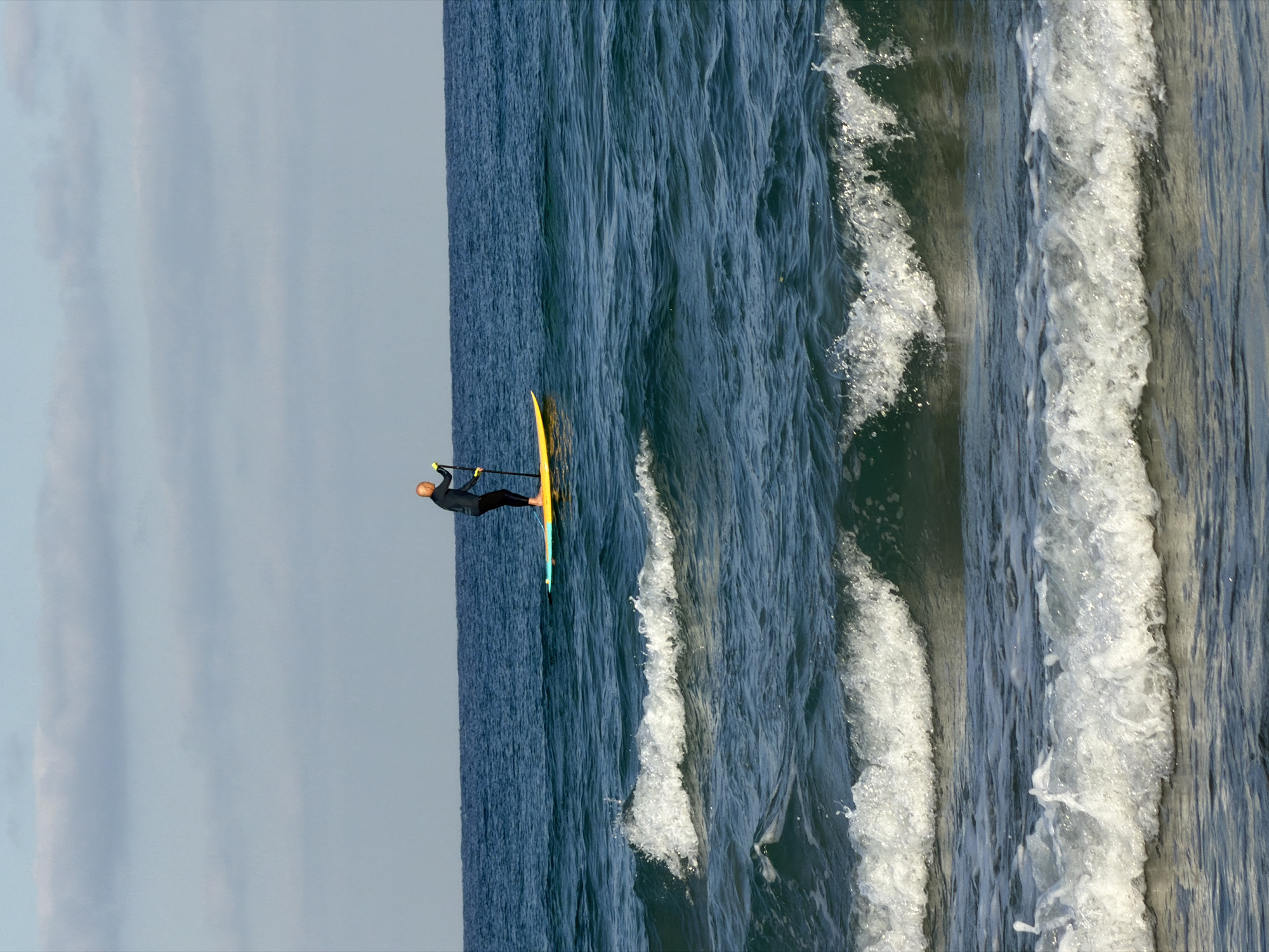 A person wearing a dark wetsuit stands on a yellow and blue paddleboard in the ocean. The person holds a paddle, which is partially submerged in the water. The paddleboard is positioned to the right of the image's vertical center. The ocean is a deep blue, with white-capped waves in the foreground. The sky above is a pale blue with scattered white clouds. The scene is likely captured at a beach in Bat Yam, Israel, given the characteristic color of the water and the clear sky.