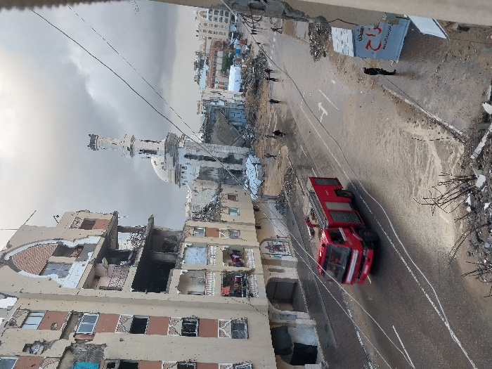 An elevated view captures a street scene in Khan Yunis, Palestinian Territory, under an overcast sky. The foreground shows debris and exposed rebar, suggesting structural damage to the immediate vicinity. A prominent multi-story building on the left exhibits severe damage, with multiple floors exposed, walls partially collapsed, and numerous impact holes. Some windows are missing, and a balcony on a lower floor shows clothes hanging. In the middle ground, a paved road, wet with standing water and puddles, features white lane markings. A red fire truck travels from left to right on this road.
Beyond the road, a white mosque with a distinct minaret and dome is visible, also appearing to have sustained damage, particularly around its base. Several individuals are present on the street, walking amidst scattered rubble and debris. One person stands near a partially damaged sign displaying Arabic text on the right side of the road. Utility lines traverse the upper and middle sections of the image. Further into the background, additional damaged structures and what appear to be temporary shelters or tents are discernible. The overall environment indicates extensive destruction and displacement.