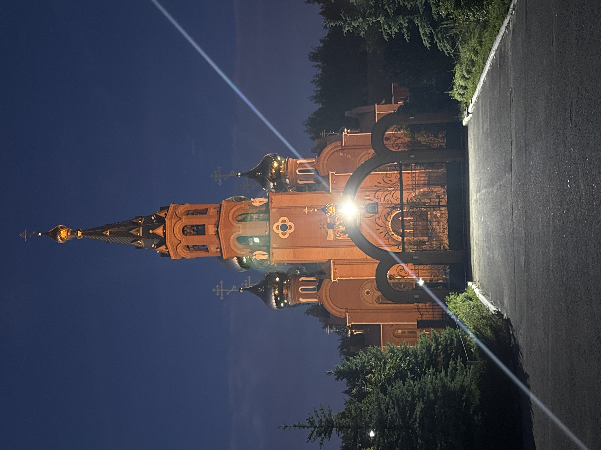 The image, rotated 90 degrees clockwise from its original orientation, depicts an illuminated Eastern Orthodox church at night in Chaikovsky, Russia. The impressive brick-colored building features a tall, dark spire crowned with a golden cross, and multiple onion-shaped cupolas, some of which are gilded and reflect light brightly. The church facade exhibits intricate architectural details, including arched windows and decorative patterns.
To the right of the main building, a grand dark metal gate with elaborate scrollwork and circular motifs is visible. A bright light source, likely a floodlight or streetlamp, is positioned near this gate, illuminating the church and creating a prominent diagonal lens flare that cuts across the dark, clear night sky.
Lush green trees are situated at the bottom and top right edges of the frame, providing a natural setting. A paved path or road runs alongside the right side of the image, leading towards the church gate. The time of day is clearly nighttime, evident from the deep blue sky and the strategic artificial lighting highlighting the church's features. There are no people visible in the scene, and no discernible text. The architectural style strongly suggests an Eastern Orthodox religious site.