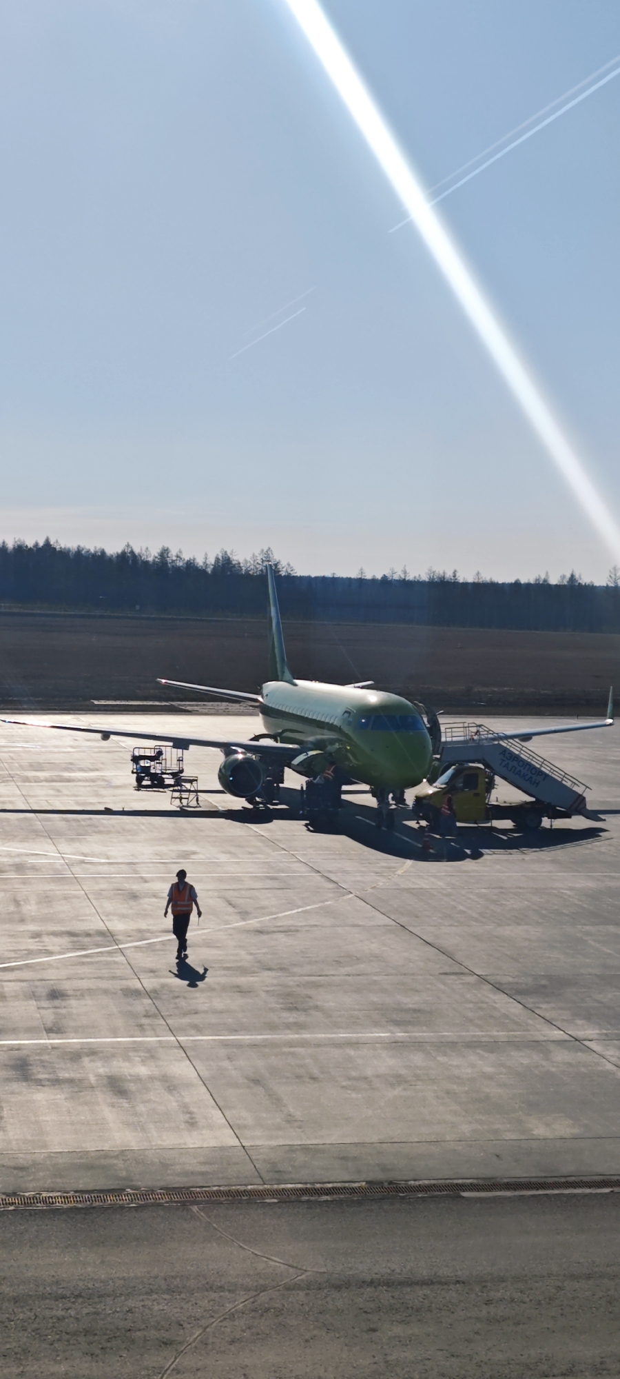 A green and silver Embraer E-Jet aircraft sits on the tarmac at Аэропорт Талакан (Talakan Airport) in Талакан, Russia, under a bright, sunlit sky. A person wearing an orange vest walks across the tarmac toward the plane. Ground support equipment, including a baggage cart and passenger stairs attached to a vehicle, are positioned near the aircraft. Trees are visible in the background, suggesting the airport is located near a forested area. A contrail is visible in the sky. The tarmac is a gray concrete surface.