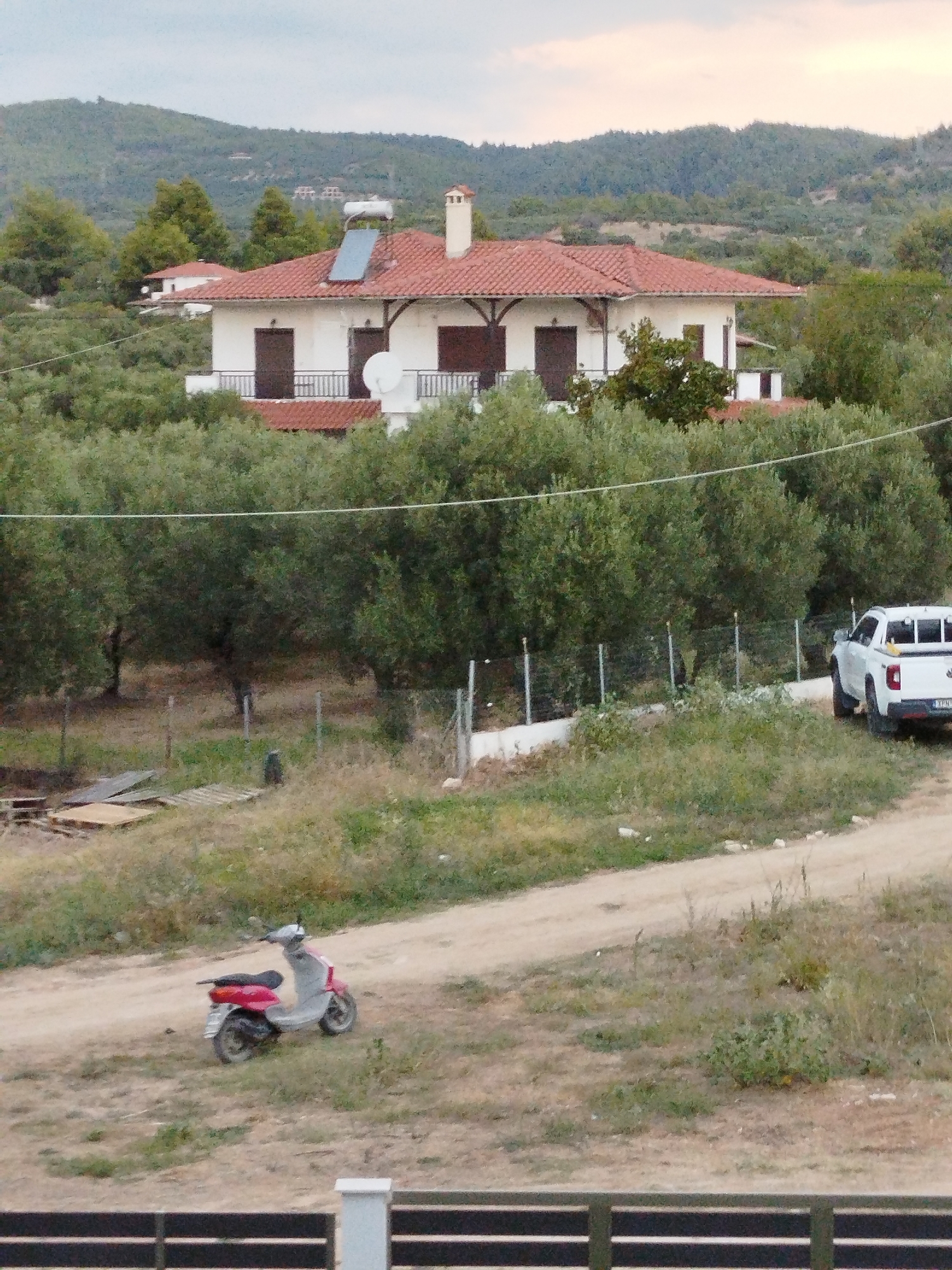 The image depicts a rural scene in the Sithonia Municipal Unit, Greece. In the background, a two-story white house with a red tile roof is visible, surrounded by trees. The house has several windows and balconies, and a solar panel is installed on the roof. In the middle ground, a white pickup truck is parked on a dirt road. A pink and red scooter is parked on the side of the road in the foreground. The environment appears to be dry and somewhat unkempt, with dry grass and sparse vegetation. The sky is overcast.