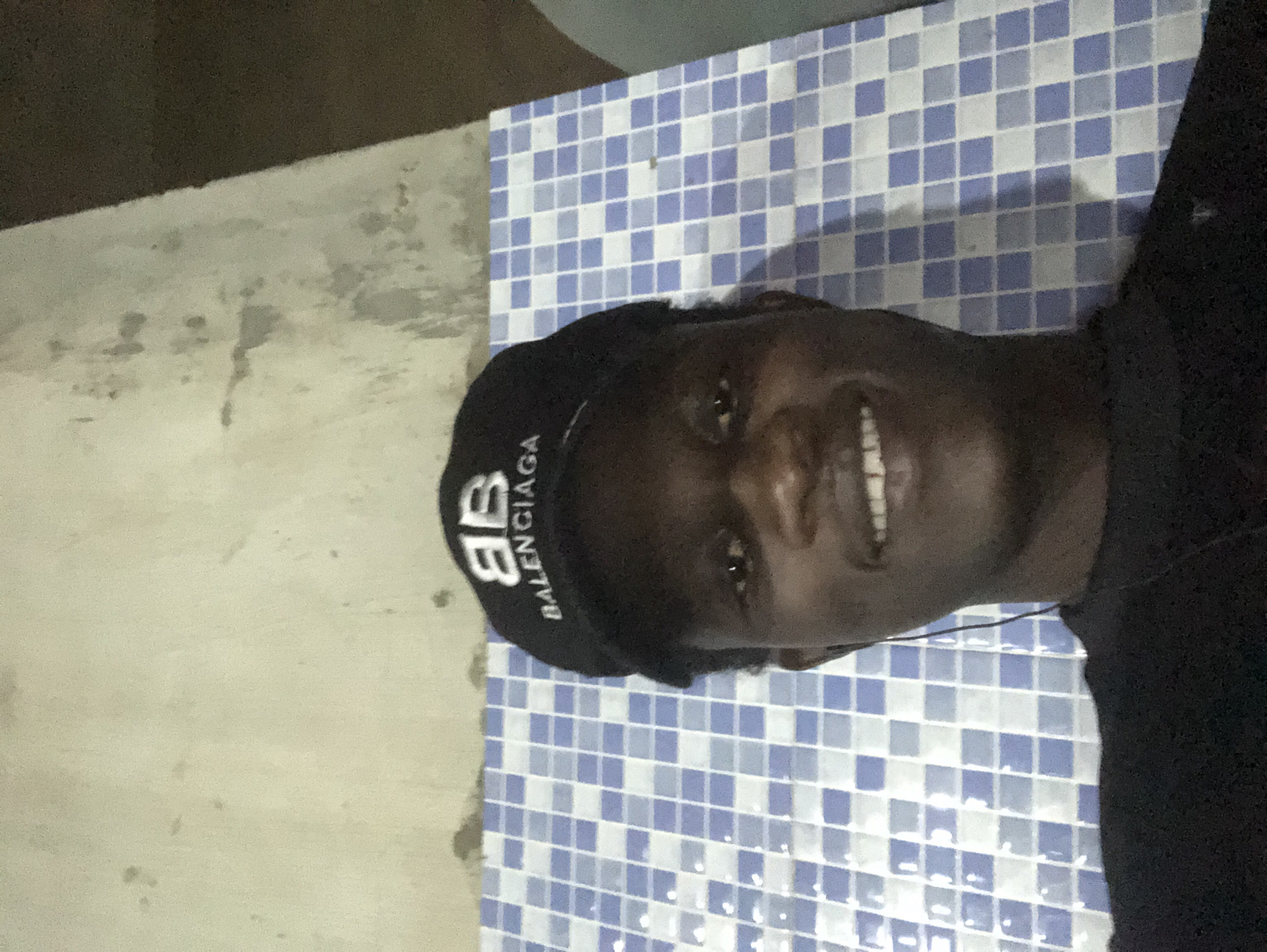 A medium shot captures a young Black male from the chest up, wearing a black baseball cap with the Balenciaga logo. He is smiling, and his eyes are open. He appears to be wearing earbuds, with a wire visible near his left ear. The background consists of a tiled surface with a blue and white checkerboard pattern, and a rough, light-colored wall above it. The lighting suggests an indoor environment, possibly at night. There are no other people or identifiable objects in the scene. The image does not provide any specific information about the location within Ado, Nigeria.
