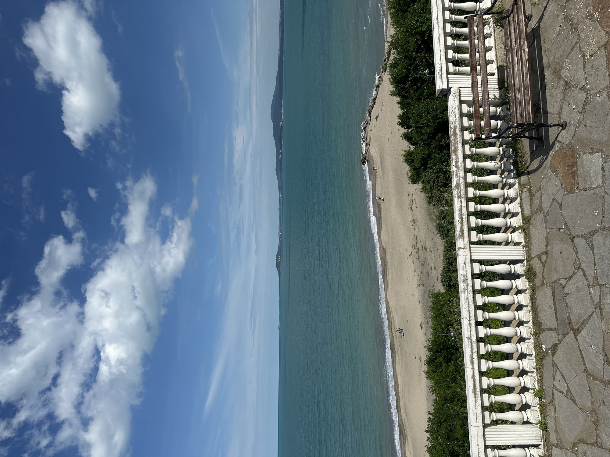 An outdoor, eye-level shot captures a scenic view of the Black Sea from a promenade in Burgas, Bulgaria. In the foreground, a stone-paved area is bordered by a white balustrade with ornate pillars and a weathered wooden bench. Beyond the balustrade, a sandy beach stretches across the mid-ground, with a few scattered people visible. The turquoise waters of the sea meet the shore, forming gentle waves. In the distance, a hazy coastline with rolling hills and a mountain dominates the horizon under a bright blue sky dotted with white, fluffy clouds. The scene evokes a sense of tranquility and natural beauty.