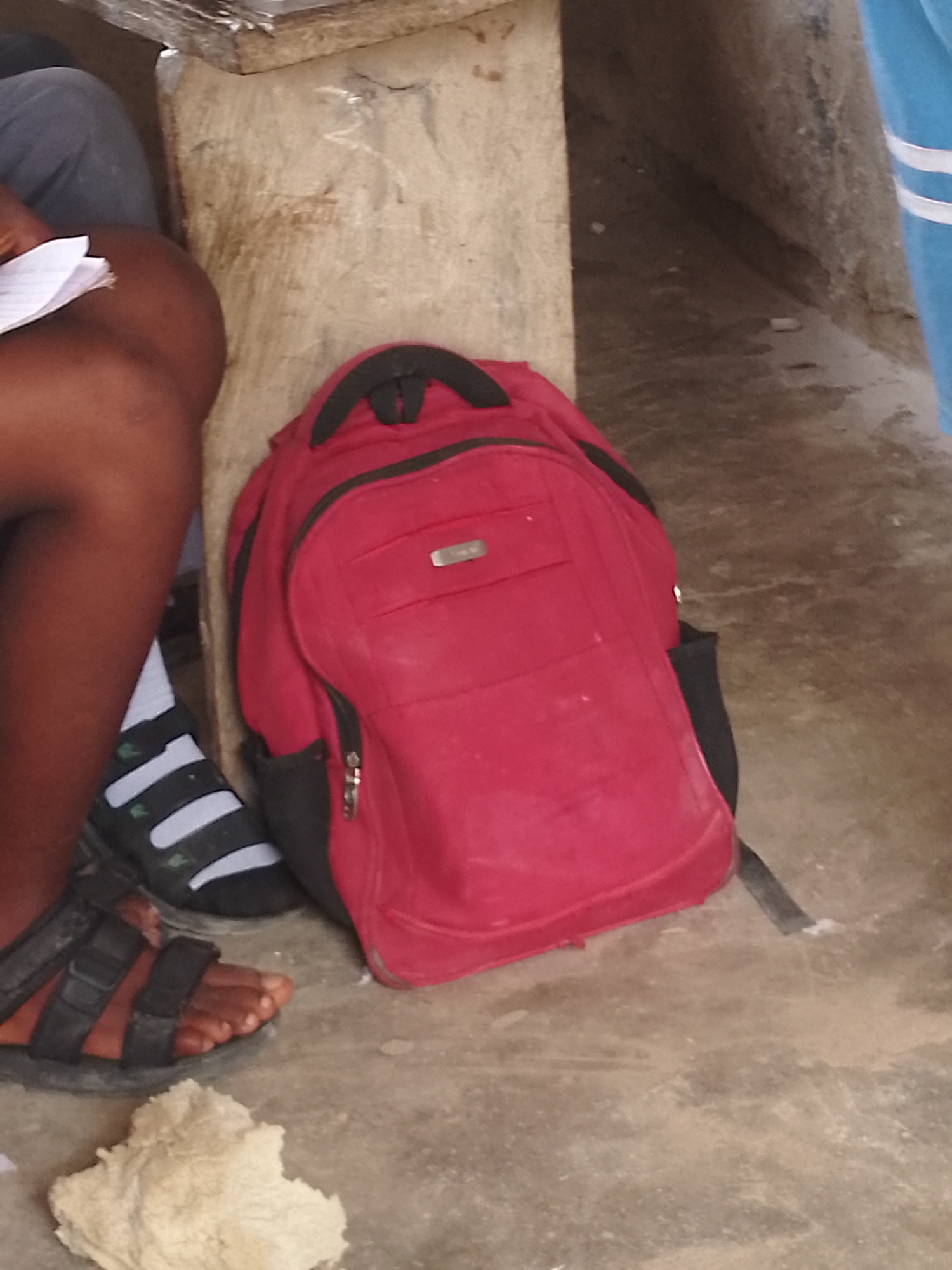 A close-up, low-angle shot shows a red backpack with black trim resting on a concrete floor. The backpack has zippers and pockets. To the left of the backpack, a person's bare foot is visible wearing a black sandal with white stripes. The person's leg, which appears to be dark-skinned, is also visible. They are wearing dark pants. A piece of white paper is held by a hand that is out of frame. To the right of the backpack, the leg of a person wearing blue pants with white stripes is visible. The background consists of a concrete wall and what appears to be a wooden structure above it. A lumpy, pale object, possibly food, is on the floor in the foreground. The lighting is natural, casting shadows on the floor.