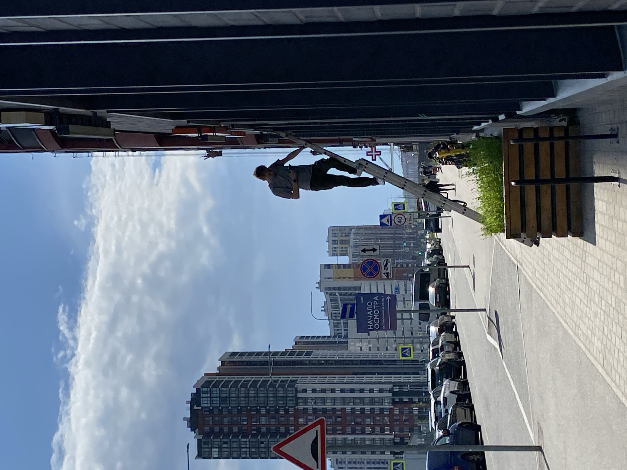 The image displays a daytime urban scene under a clear blue sky with some scattered white clouds. In the foreground, a man is positioned on a tall, silver extension ladder, which leans against the dark, multi-storied facade of a modern building. Dressed in a grey t-shirt and dark trousers, the man appears to be engaged in maintenance or installation work, looking upwards and reaching with his right hand.
To the right of the ladder, a patterned, light-colored sidewalk extends into the background, bordering a planter box filled with green shrubbery. Beyond this, a street is lined with several parked cars. The background is dominated by numerous high-rise residential or commercial buildings, showcasing contemporary architectural designs.
Various traffic and informational signs are visible along the street. These include a circular speed limit sign displaying "40", a blue rectangular sign with an arrow and the Cyrillic text "НАЧАЛО ОСМОТРА" (Start Inspection), a red triangular warning sign for a speed bump, and other regulatory and directional signs, such as a pedestrian crossing symbol and a no-stopping/parking sign. A distinct red cross symbol on a white square is also visible on the building facade, potentially indicating a medical facility or pharmacy. The overall setting portrays an active city environment, consistent with an urban area in Saint Petersburg, Russia, as suggested by the Cyrillic text.