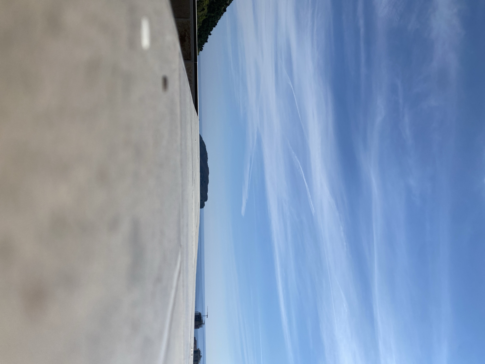 The image shows a view from what appears to be a balcony or raised platform in Budva, Montenegro. To the left is a light-colored, smooth, possibly concrete or stone surface, likely the balcony's edge. Beyond this surface is a body of water, probably the Adriatic Sea, extending to the horizon. An island or a promontory covered in dark green vegetation is visible in the middle distance. In the distance, what seems to be construction structures or buildings are on the water. The sky occupies the majority of the frame and is a clear blue with streaks of cirrus clouds, possibly indicating contrails from airplanes.