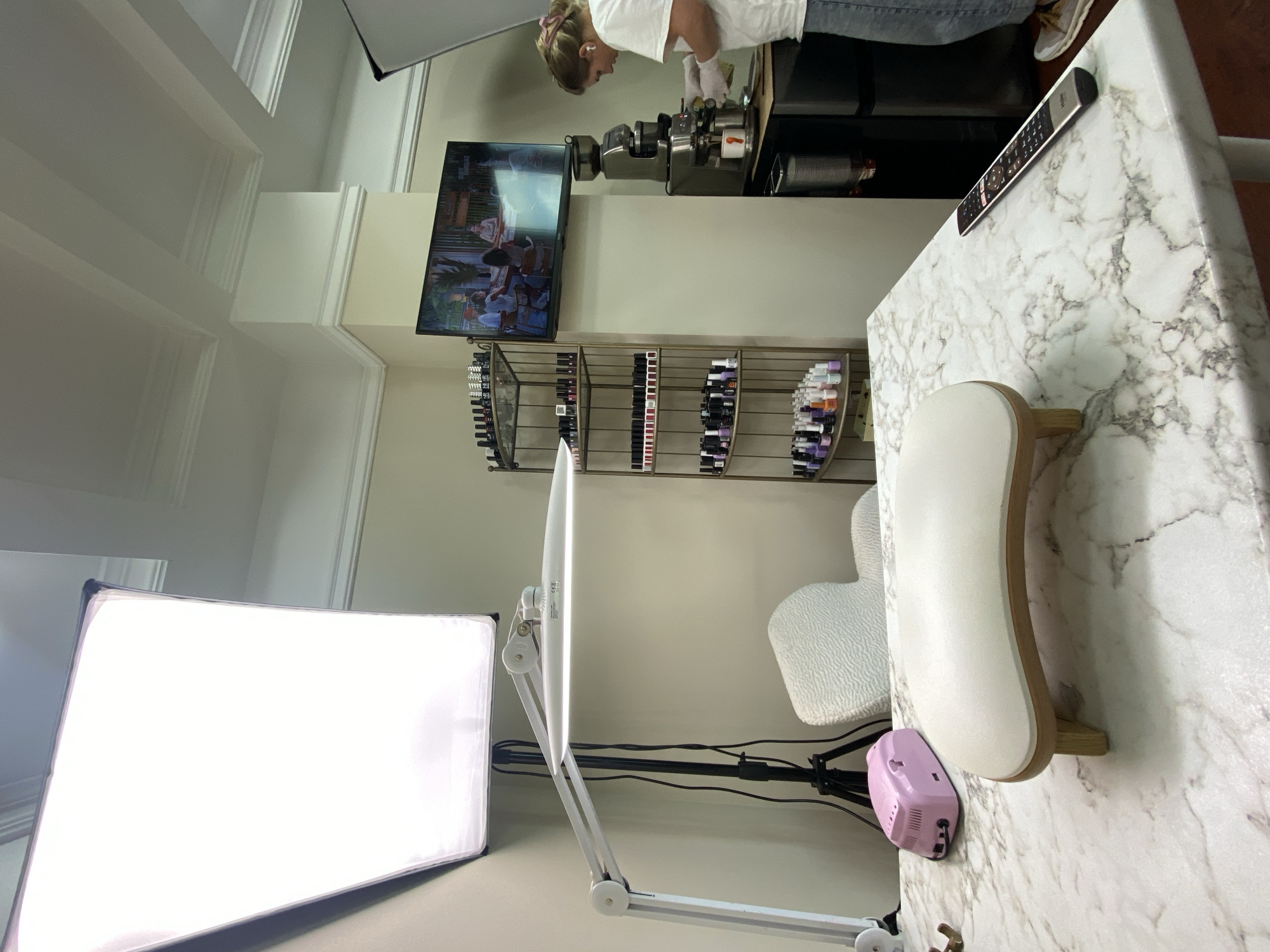 The image depicts an indoor setting, potentially a nail salon. A person wearing a white shirt and jeans is working at a station with various tools and equipment. A television screen shows a video playing on the wall behind the station. Shelves holding nail polish bottles are mounted on the wall. The foreground contains a brightly lit rectangular panel, likely a lighting tool. A white manicure table with a marble-like surface is in the foreground. A hand rest sits on the table, along with a pink object, possibly a nail drill. A white chair sits at the table. A remote control rests on the table. There is no explicit indication that this is in Sochi, Russia.