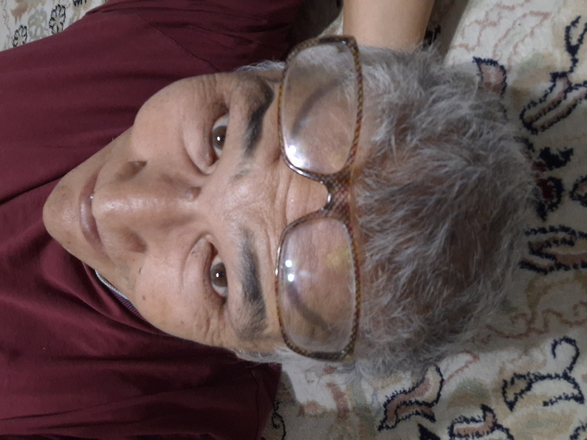 The image is a close-up, upside-down selfie of an individual wearing a dark red shirt. The person has gray hair, is wearing glasses with a tortoiseshell pattern on the rims, and appears to be lying down on a patterned carpet. The background shows a portion of a maroon-colored garment and a cream-colored carpet with intricate floral and geometric patterns in muted colors like pink, beige, and dark blue. The lighting suggests an indoor setting. There is no visible information to indicate the location is in Almaty, Kazakhstan.