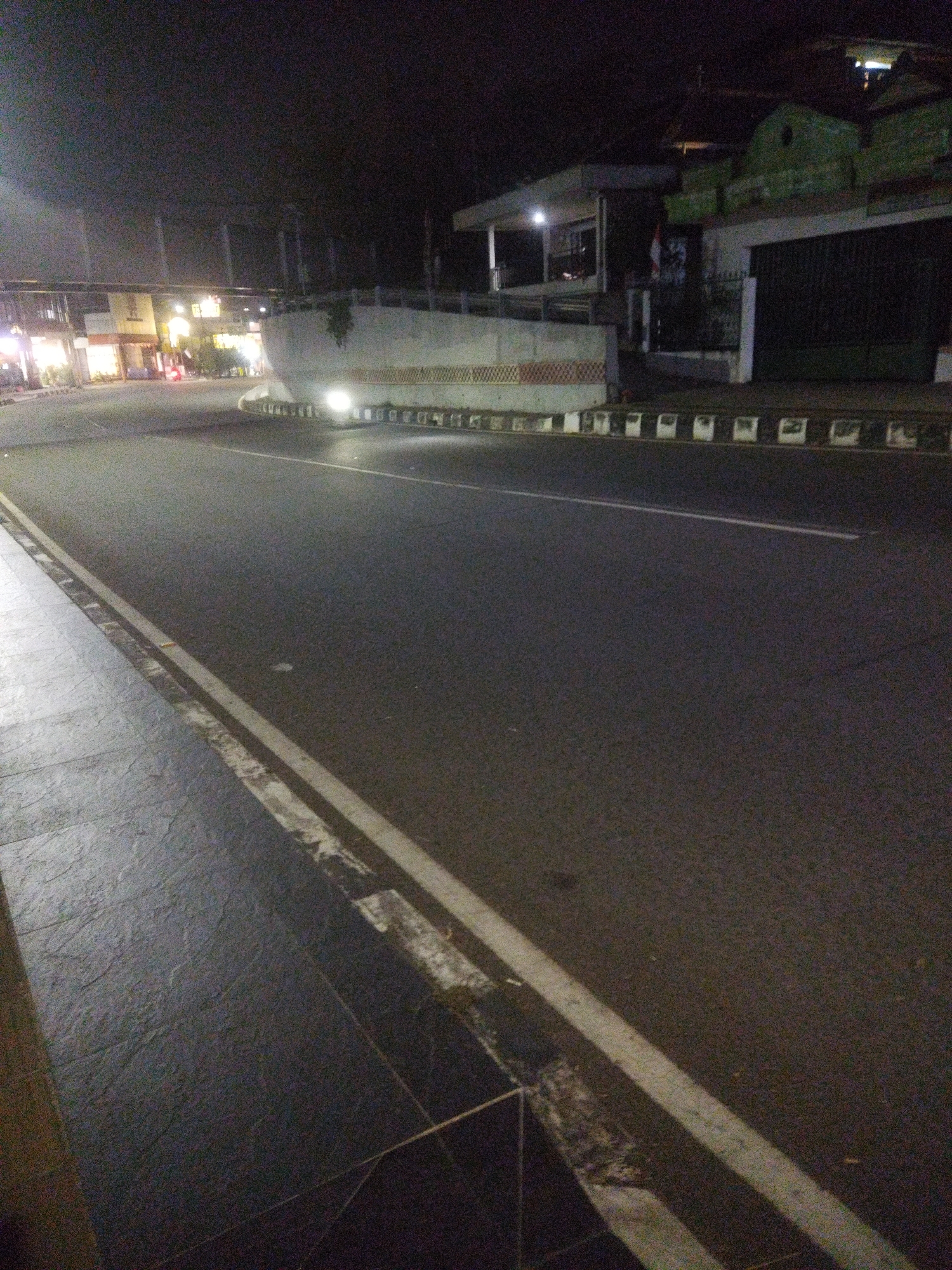 This is a night-time photograph of a street in Wonogiri, Indonesia. The foreground shows a paved sidewalk next to a road with a double yellow line marking. In the background, there are buildings with some lights on, indicating commercial activity. A curved concrete barrier with patterned detailing is visible in the middle ground, and there are lights from vehicles illuminating the road and the barrier. The sky is dark, typical of nighttime. The overall scene depicts a typical urban street environment after dark.