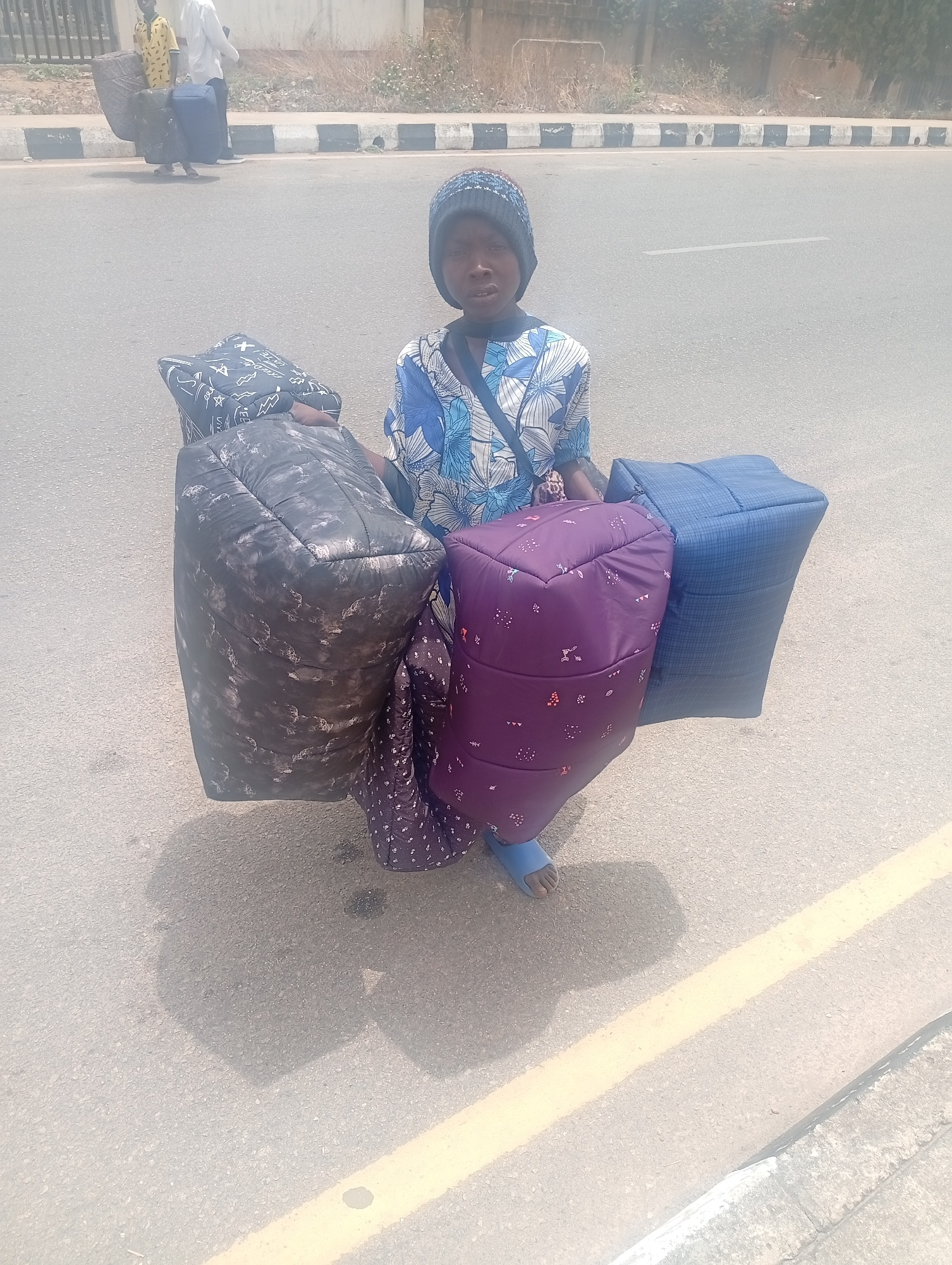 A young person in Jos, Nigeria stands on a gray asphalt road, bearing four stuffed, patterned bags in front of them. They are wearing a blue and white floral shirt, a dark knit cap, and blue sandals. The road has a yellow line near the right curb. In the background, two other people are walking along the road, also carrying bags. A low concrete barrier runs along the edge of the road. A building is visible in the background.