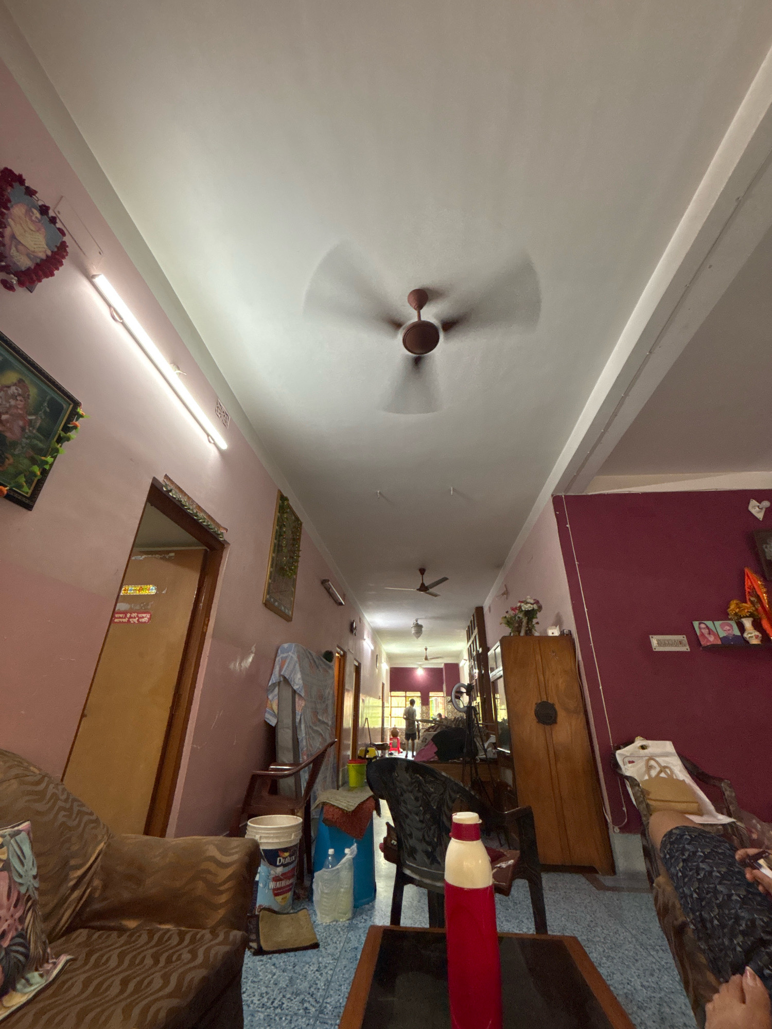 The image shows an interior hallway with a ceiling fan in motion. The walls are painted pink on the left and a dark reddish-purple on the right. On the left wall, there are framed pictures and a door. On the right wall, there are more framed pictures and a wooden cabinet. In the foreground, there is a sofa and a table with a red and white water bottle. In the middle ground, there are chairs and buckets. The floor is tiled. The lighting appears to be from overhead fixtures and a long fluorescent light on the left wall. The scene is cluttered with various household items. The context of Bally, India is not discernible from the visual information alone.