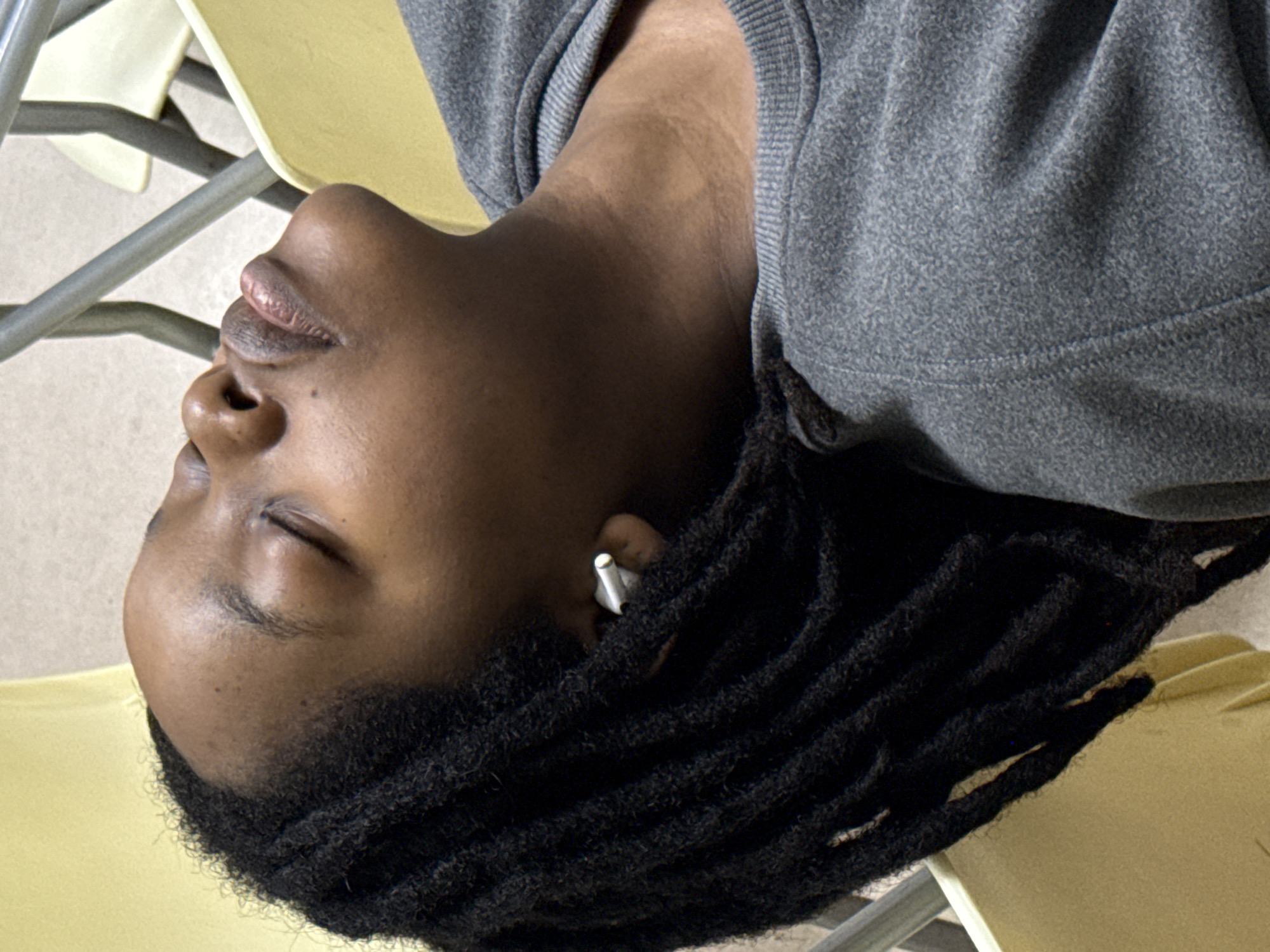 A close-up, rotated image shows a person with dark skin lying on yellow chairs. The person is wearing a gray top and has black dreadlocks. An earbud is visible in the person's ear. The background is a neutral color, likely a wall or floor. There is no indication of the location being Yuzhong District, China.