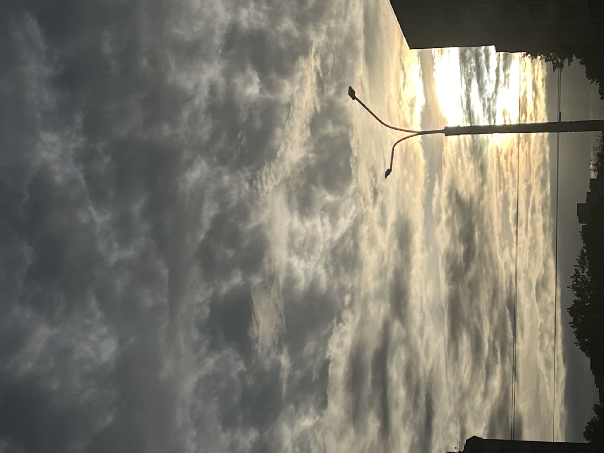 The image captures a dramatic sky filled with a mix of dark and bright clouds, suggesting either the approach or departure of a storm, or perhaps a sunset or sunrise. The sun is a prominent feature, a bright, diffused light source peeking through gaps in the clouds, casting a golden glow on the scene. A modern-looking street lamp with a distinctive orange-red arm and two lamp heads stands tall in the foreground, its structure silhouetted against the illuminated sky.
In the background, the dark outlines of buildings and some silhouetted trees hint at an urban or suburban setting. The clouds themselves are varied in texture and color, with large swathes of dark grey appearing heavy and dense, contrasting with streaks of brilliant white and yellow where the sunlight breaks through. The overall mood evoked is one of atmospheric intensity and natural beauty. The orientation of the image suggests it might have been taken in portrait mode, or possibly rotated. There is no visible text in the image.