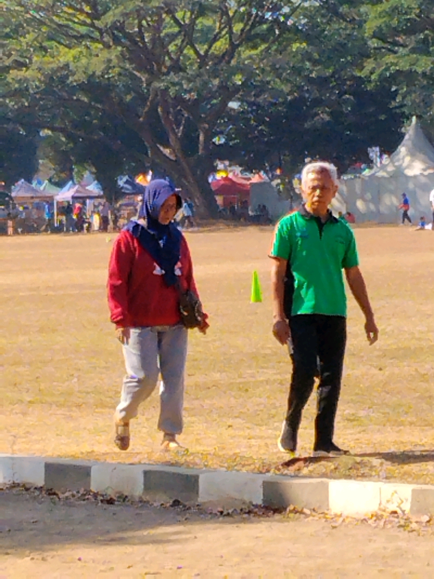 In Malang, Indonesia, two individuals walk across a dry, grassy field under a bright sky. On the left, a woman wearing a navy blue hijab and a red jacket walks with a dark bag slung over her shoulder, paired with light gray pants and sandals. To her right, a man with gray hair, dressed in a green polo shirt and black pants with white-striped socks, walks with a determined stride. In the background, tents are set up, suggesting an outdoor event or market. A green traffic cone stands prominently in the field, and a low concrete barrier runs along the foreground. The overall scene is one of daytime activity in an open, outdoor setting.