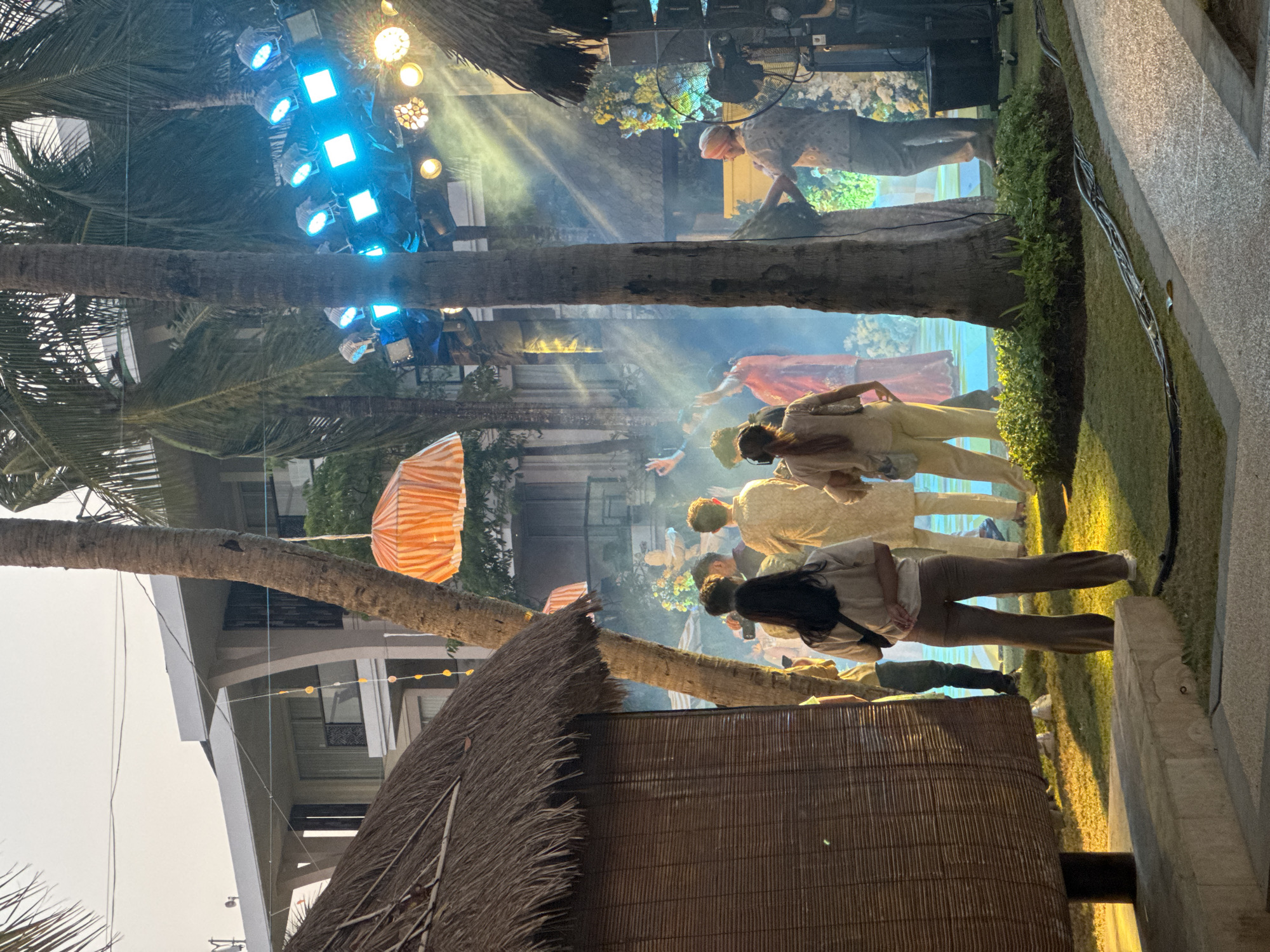 The image depicts an outdoor event in Kuta Selatan, Indonesia, likely a cultural performance or celebration. In the foreground, a thatched-roof hut and palm trees frame the scene. Several people are gathered, some facing a stage illuminated by bright blue lights and spotlights that create dramatic beams through the mist. Performers in traditional attire are visible on the stage. The atmosphere suggests an evening event, possibly a wedding reception or a festival, with decorative elements like orange and white umbrellas hanging above. The background shows a modern building, typical of resorts or hotels found in Kuta Selatan.