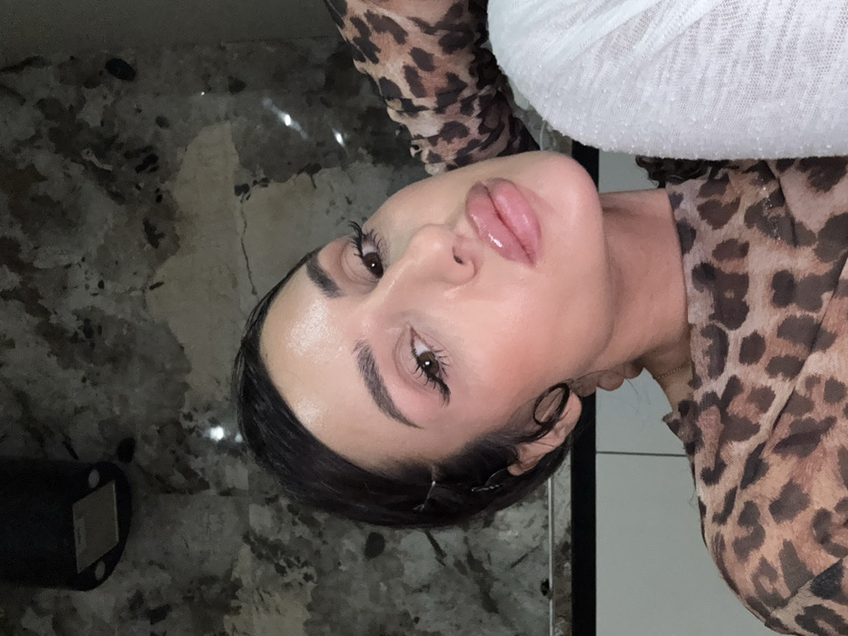 A person's face is centered in the image, tilted slightly. They have dark hair styled back from their face. The individual has full lips and appears to be wearing makeup. They are wearing a leopard-print top, and what appears to be a white, textured garment on their shoulder. The background consists of a dark, speckled surface and a white wall with black trim.