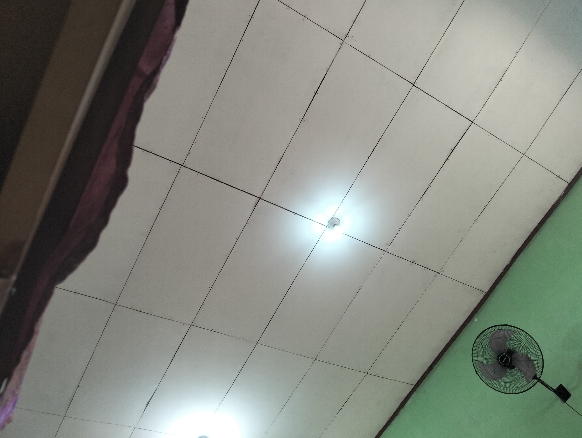 The image is taken from a low angle, looking upwards towards a ceiling. The ceiling appears to be made of white square tiles with dark lines separating them. Two bright lights are visible on the ceiling, one in the upper middle and another in the lower left corner, creating a halo effect. To the left of the ceiling, a dark, textured material, possibly fabric, hangs down. To the right, the ceiling transitions to a green wall, marked by a dark brown border. A black oscillating fan with a circular grille is mounted on the green wall in the lower right portion of the image. The scene depicts an indoor environment with a focus on the ceiling, wall, and fan. It is not possible to determine the specific location within Banjar Agung, Indonesia, from the visual information provided.