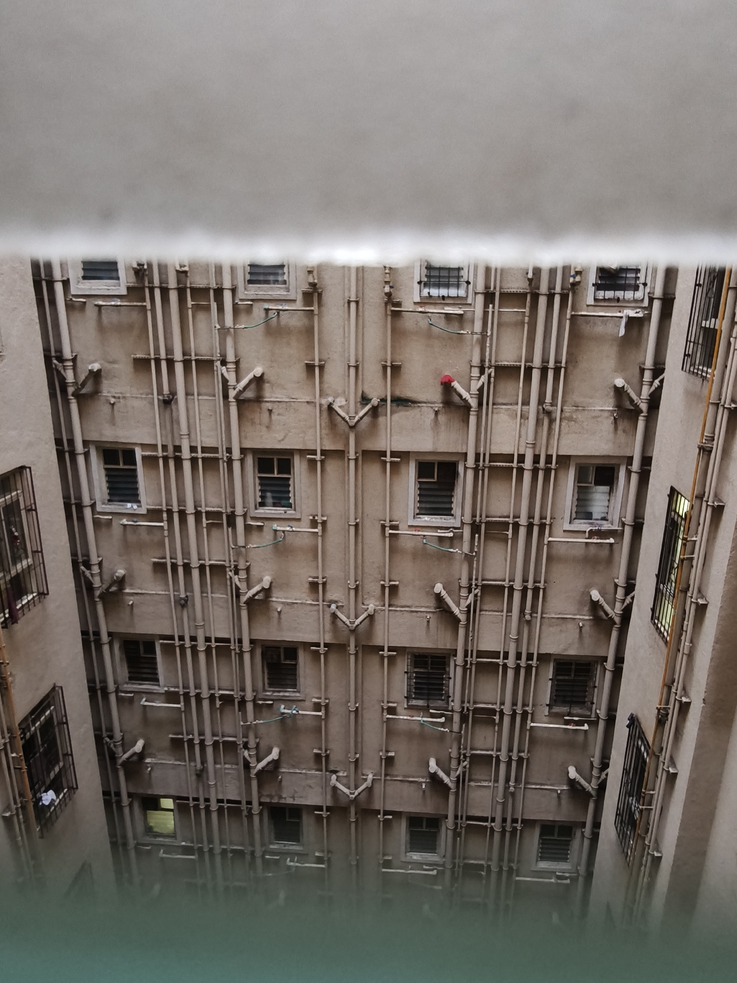 The image shows an aerial view looking down into a narrow courtyard between two apartment buildings. The walls of the buildings are a muted beige color and are covered in a dense network of pipes, likely for plumbing and drainage. Many windows are visible, some with bars, and some appear to be covered with louvers. There are no people or animals visible in the image. The overall impression is one of urban density and utilitarian construction. The location is identified as Bavdhan, India.