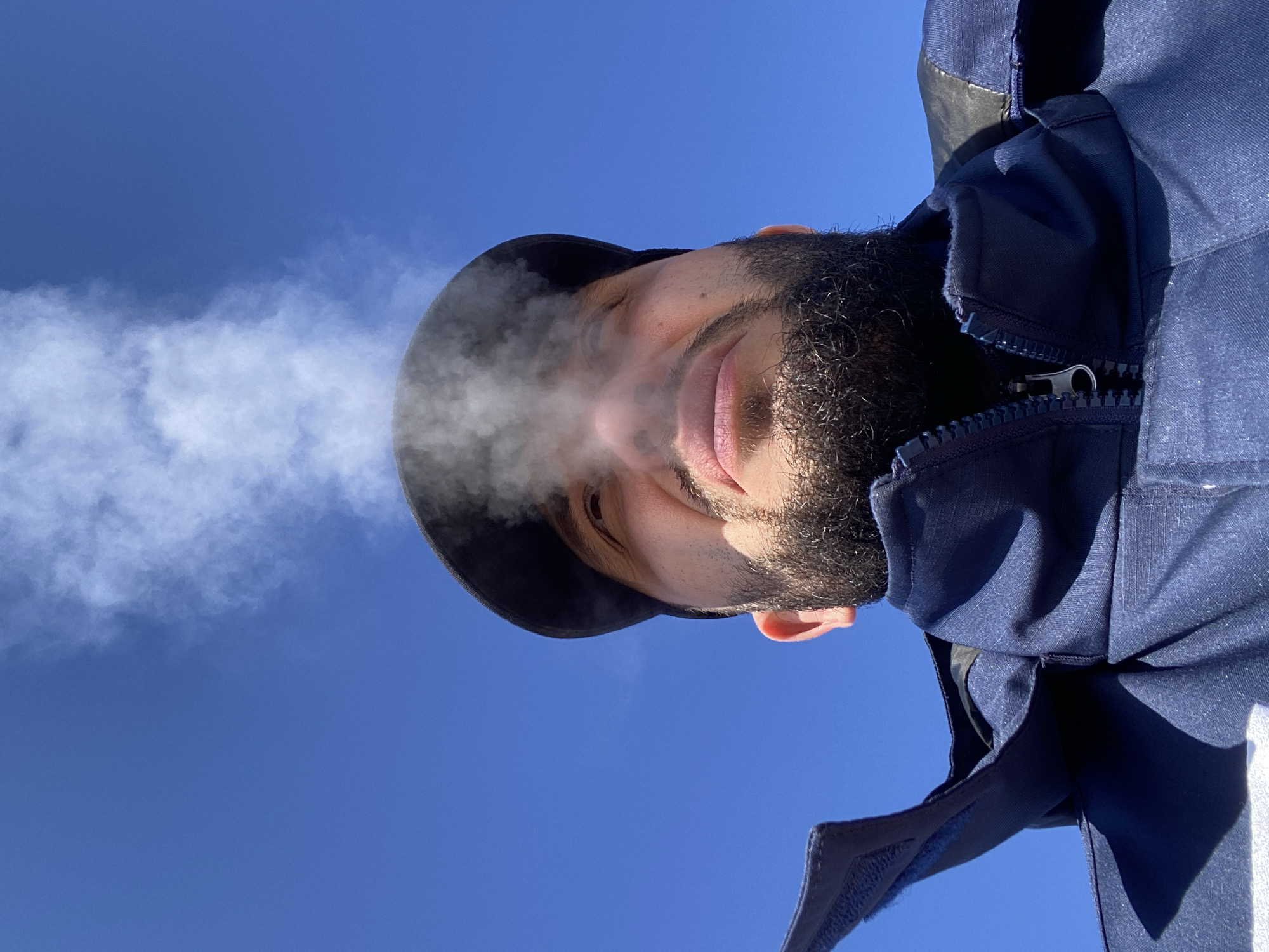 A man with a beard wearing a hat and a blue jacket is visible in the foreground of the image. White vapor or smoke is emanating from his mouth. The background is a clear, bright blue sky. The image is taken from a low angle, looking up at the man. The location is not specified, and it is impossible to determine if the image was taken in Luzhitsy, Russia, based on the provided visual information.