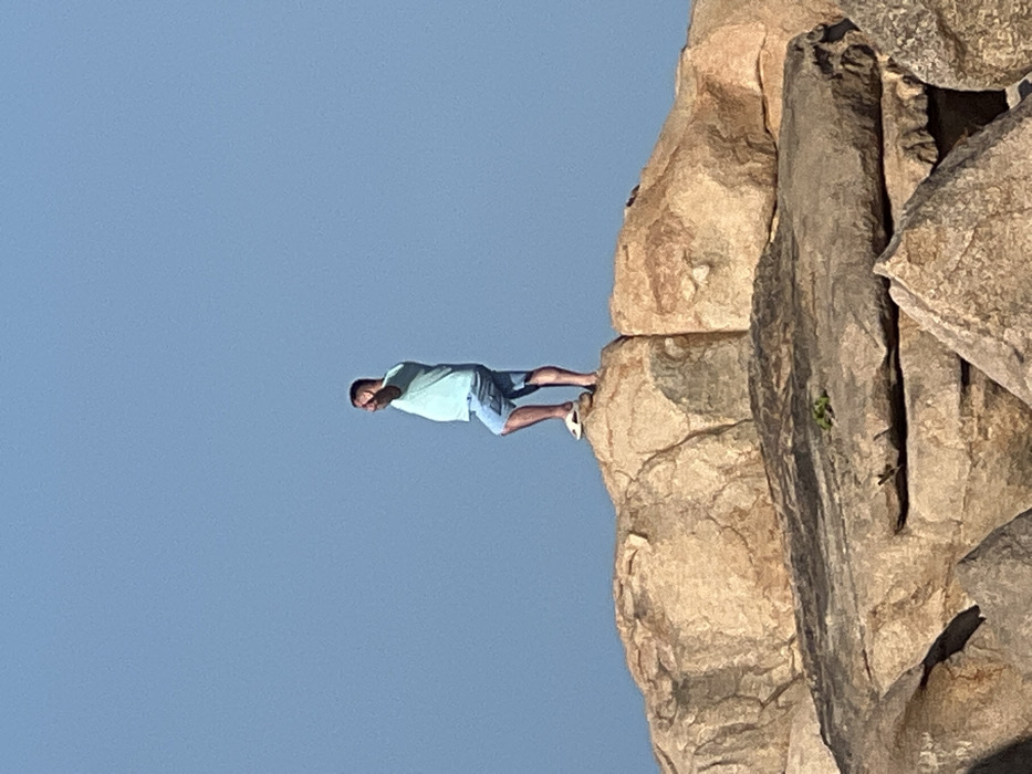 An adult male stands in profile on a large, weathered rock formation in Nha Trang, Vietnam, under a clear, pale blue sky. He wears a light blue short-sleeved t-shirt, light blue denim shorts, and white flip-flops. His right arm is raised, with his hand open and fingers splayed, pointed forward. The man's left leg is slightly bent, and his right leg is straight. The rocks are light brown to tan with visible fissures and texture, occupying the lower two-thirds of the frame. A small patch of green vegetation is visible on a rock in the lower midground.