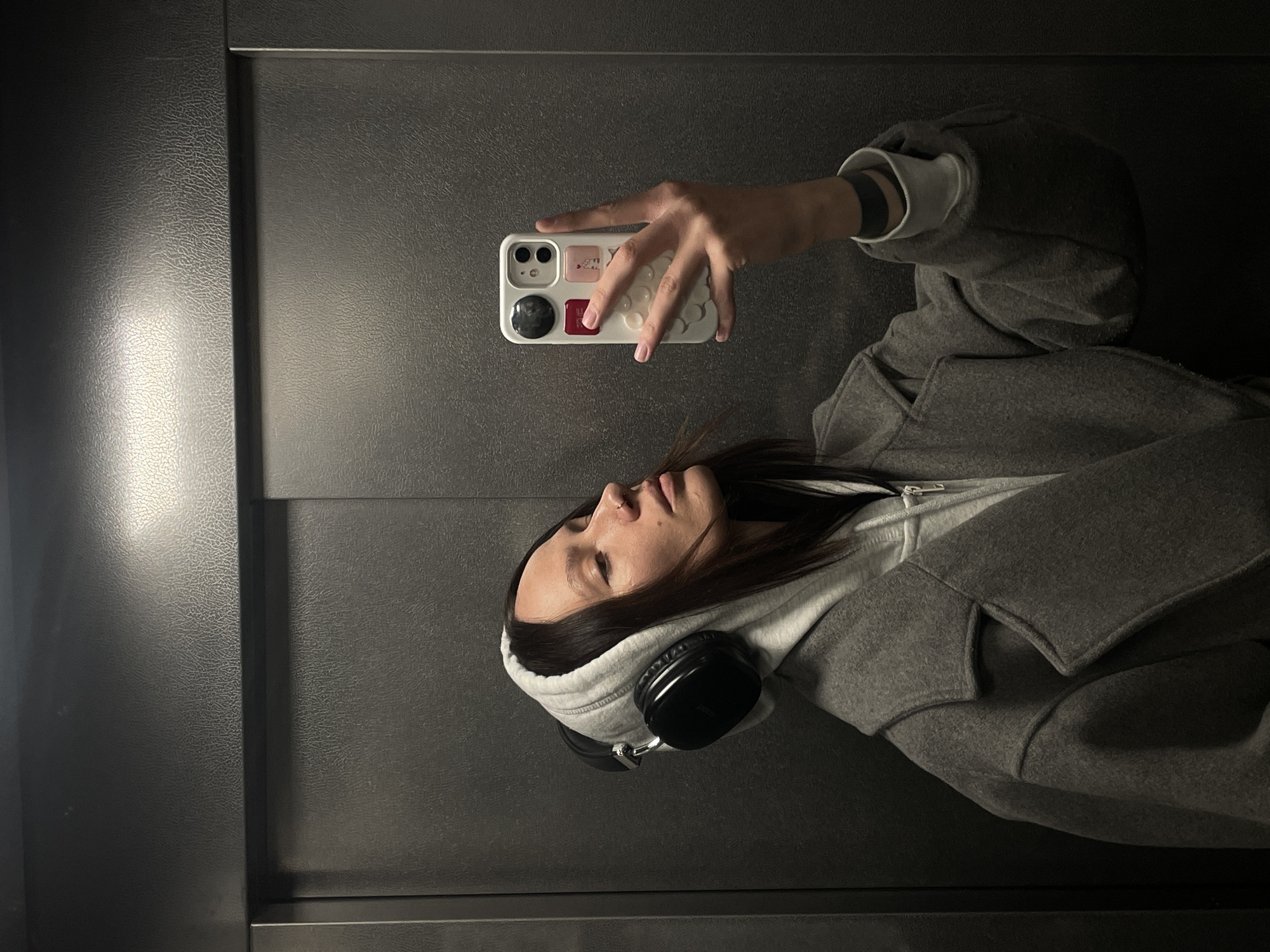 A female subject is taking a selfie in what appears to be an elevator. She is wearing a grey hoodie, a grey coat, and headphones around her neck. The phone she is holding has a white case with multi-colored accents. The elevator interior is dark grey with a textured finish. There is no definitive evidence to place the location within Izhevsk, Russia.