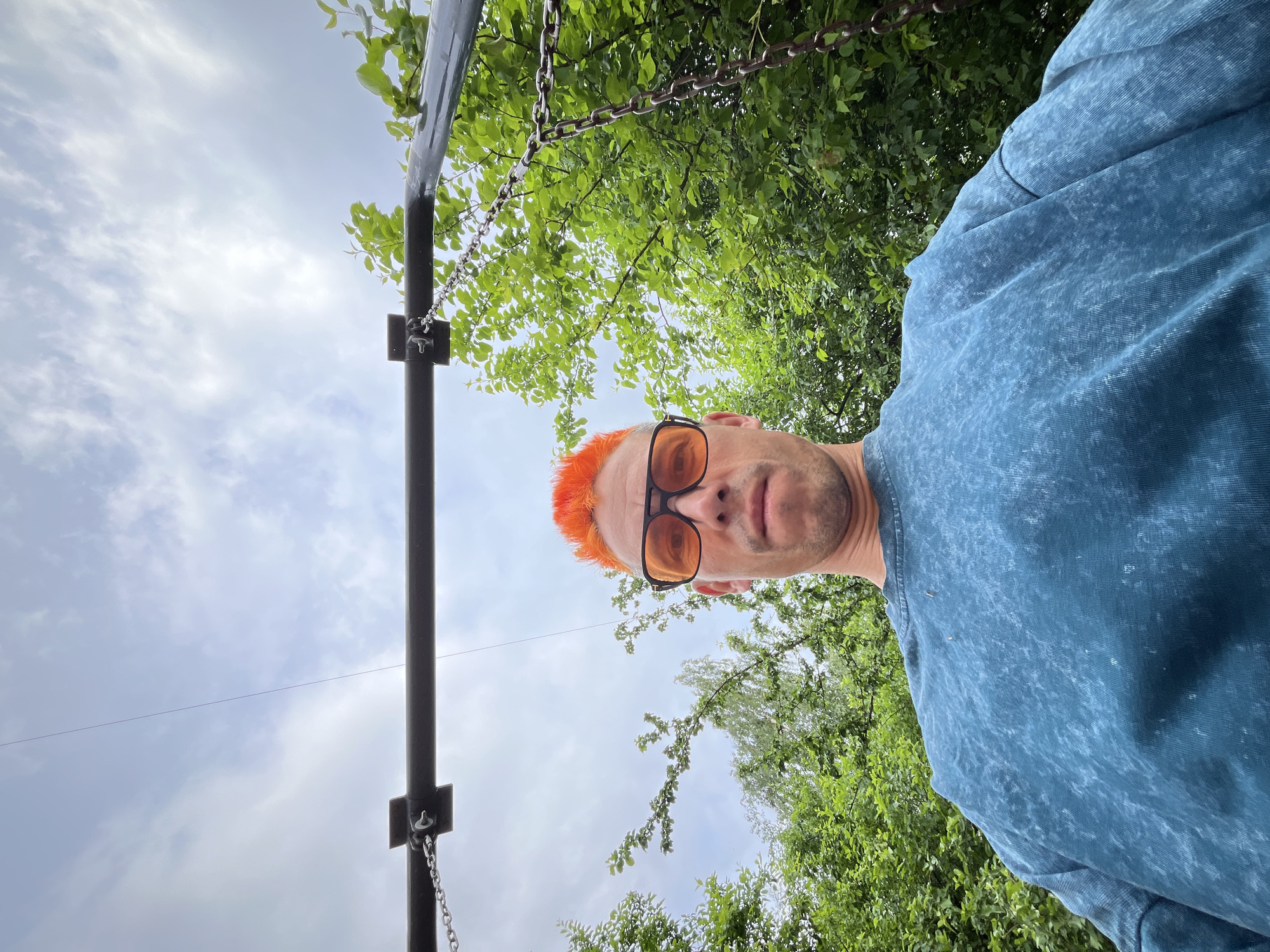 A low-angle photograph depicts a man with orange hair and orange-tinted glasses. He is wearing a blue shirt. Above and to the left of the man, a section of black metal frame with hanging chains is visible. Green foliage and sky with clouds are in the background. The location appears to be outdoors, possibly in a park-like area, based on the visible foliage. The specific location in Chaikovsky, Russia, is not identifiable from the visual data provided.