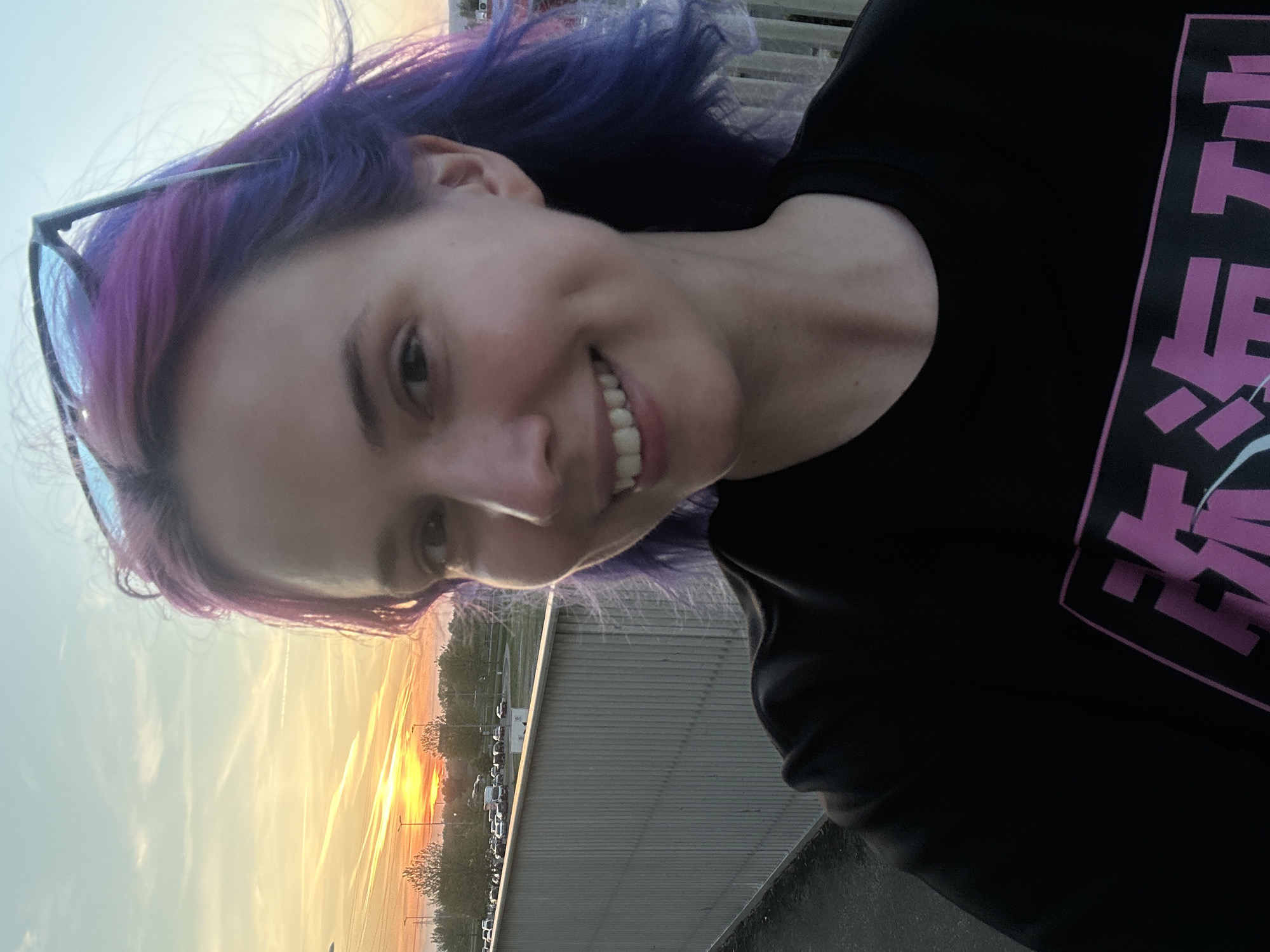A medium shot selfie of a fair-skinned woman with purple hair, wearing sunglasses perched on her head and a black t-shirt with pink graphics. The woman is smiling at the camera. The background shows a sunset over what appears to be a parking lot or cityscape. The location is unidentifiable as City of Zagreb, Croatia based on the image.