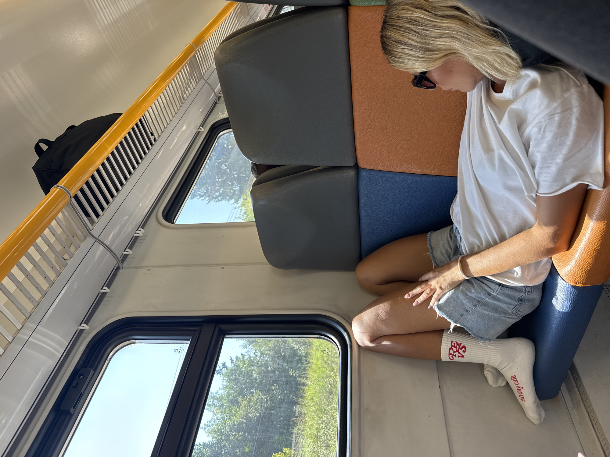 The image shows a woman resting on a train seat, with her legs extended and resting against the window. She is wearing a white t-shirt, denim shorts, and white socks with red lettering that reads "RE VS All day cafe". The train appears to be in motion, as indicated by the greenery visible outside the window, which suggests a rural or suburban landscape, possibly in or around Odintsovo, Russia. The interior of the train carriage is visible, featuring blue and orange seating, a yellow handrail, and overhead storage. A black backpack is placed on the luggage rack. The lighting suggests it is daytime, with sunlight streaming through the window.