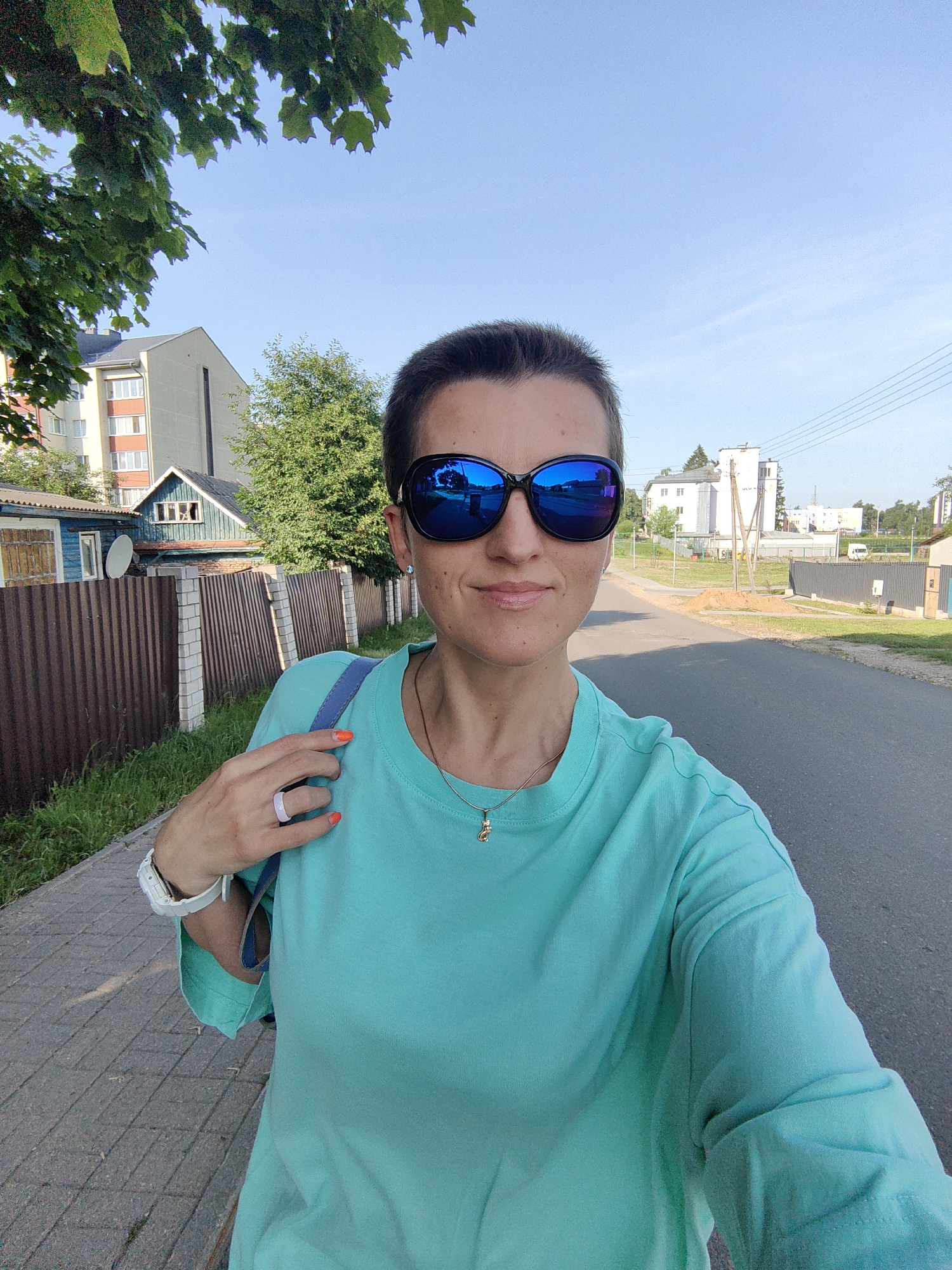 A short-haired woman wearing a turquoise t-shirt and blue sunglasses stands on a paved sidewalk. She is holding her left hand to her chest, revealing a light purple ring and bright orange nail polish. A white watch is visible on her left wrist. Behind her, a brown wooden fence with white brick posts separates the sidewalk from a grassy area and residential buildings, including a small blue house with a green roof and a larger apartment building. The scene is set on a street in the city of Fanipal, Belarus, under a clear blue sky.