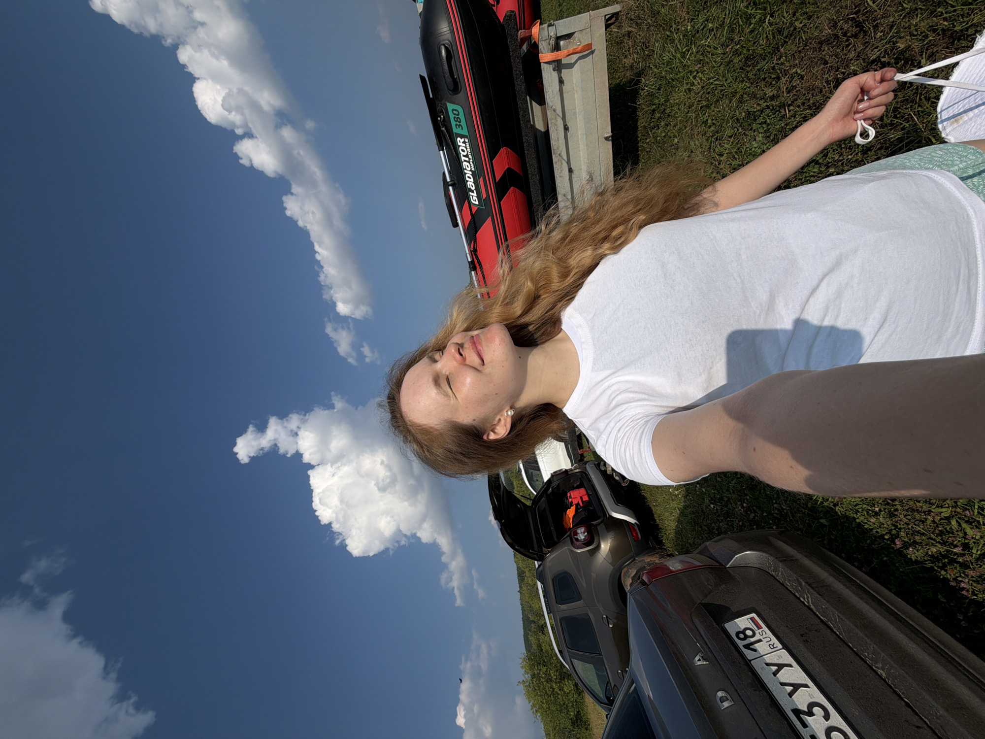 A young woman with long, wavy blonde hair is lying down, taking a selfie outdoors. She is wearing a white t-shirt and a green patterned skirt. Her eyes are closed, and she has a relaxed smile on her face, suggesting a sense of peace or enjoyment. Her left hand is raised, holding onto white cords, possibly from a hammock or tent.
In the background, a red and black inflatable boat, labeled "GLADIATOR 380", is visible on a trailer. Several cars are parked in a grassy area, with their trunks open. The scene appears to be a casual outing, perhaps a picnic or a day trip to the countryside.
The sky is a vibrant blue with scattered white clouds, indicating fair weather. The sun is casting shadows, suggesting it is daytime, likely late morning or early afternoon. The lush green grass and distant trees hint at a rural or park-like setting. The license plate on one of the cars reads "533YY 18 RUS". The overall mood is serene and summery. The location context is Krasnyi Bor, Russia.